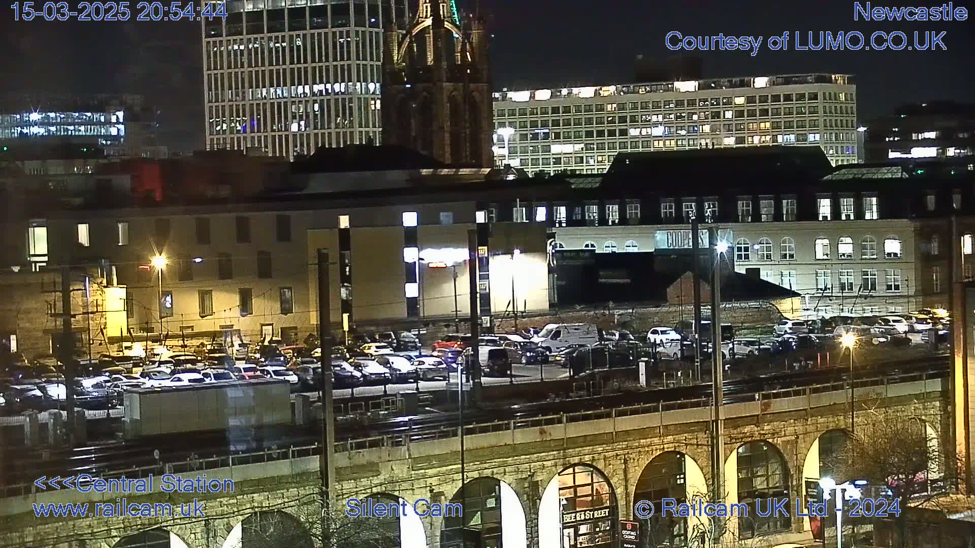 A nighttime view of a city with numerous buildings, a large parking lot filled with cars, and a stone bridge with arches.