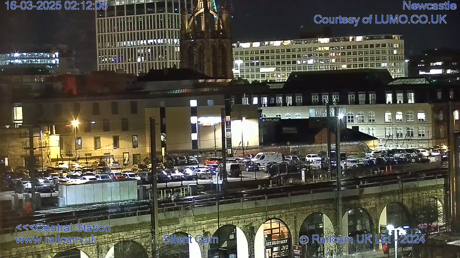 A nighttime view of a city featuring a parking lot, buildings, and a bridge, under clear skies.