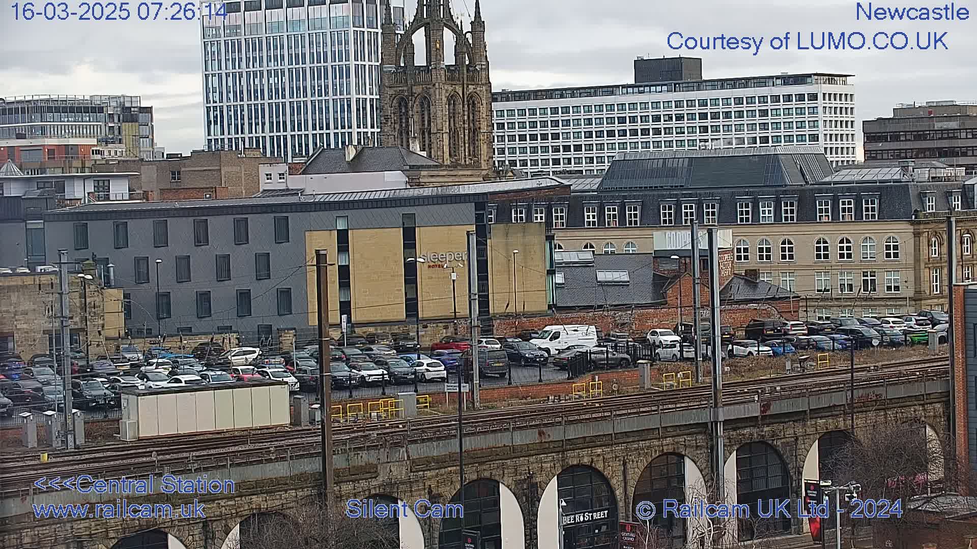 A city scene viewed from above shows a parking lot full of cars next to train tracks that pass under a stone archway, with a variety of buildings in the background under a cloudy sky.