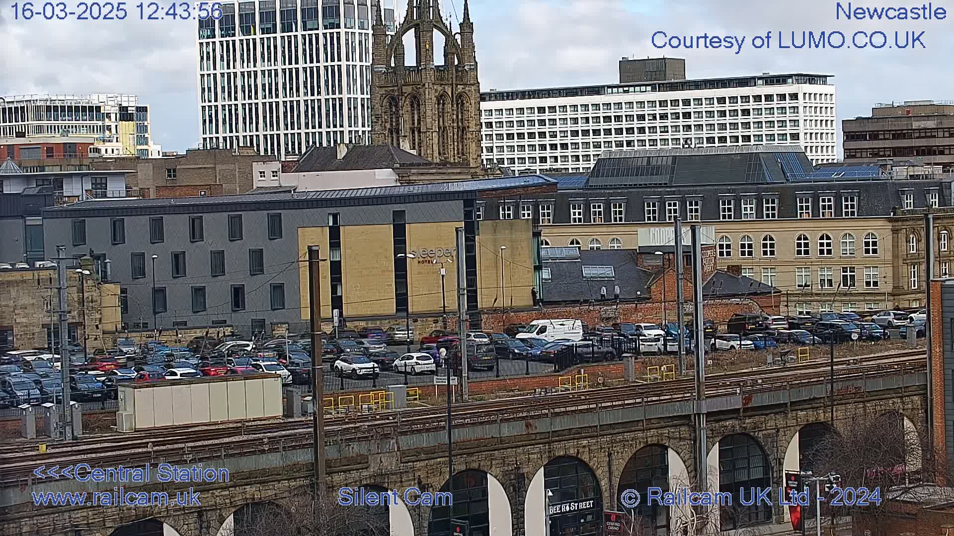 A city skyline view with a large parking lot filled with cars, situated next to a railway line and under a cloudy sky.