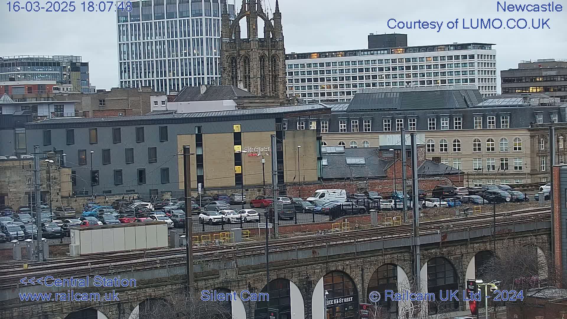 A city skyline with overcast skies, featuring a parking lot filled with cars, a train track, and various buildings including a church and a high-rise.