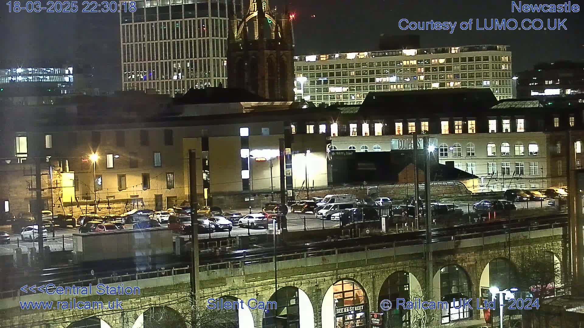 A nighttime view of a city's illuminated buildings and a parking lot, seen from above a railway viaduct under a clear, dark sky.