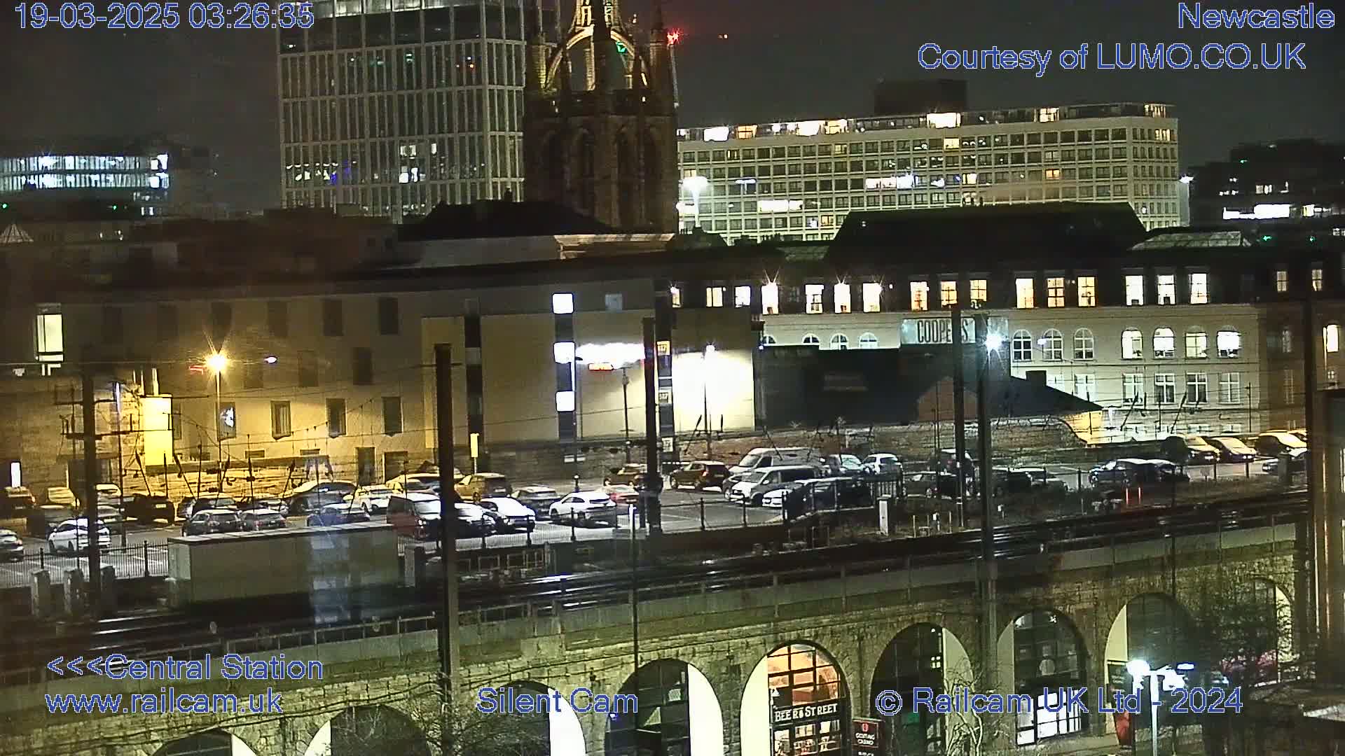 A nighttime view of a city, showing several buildings, a parking lot filled with cars, and a bridge with arched supports.