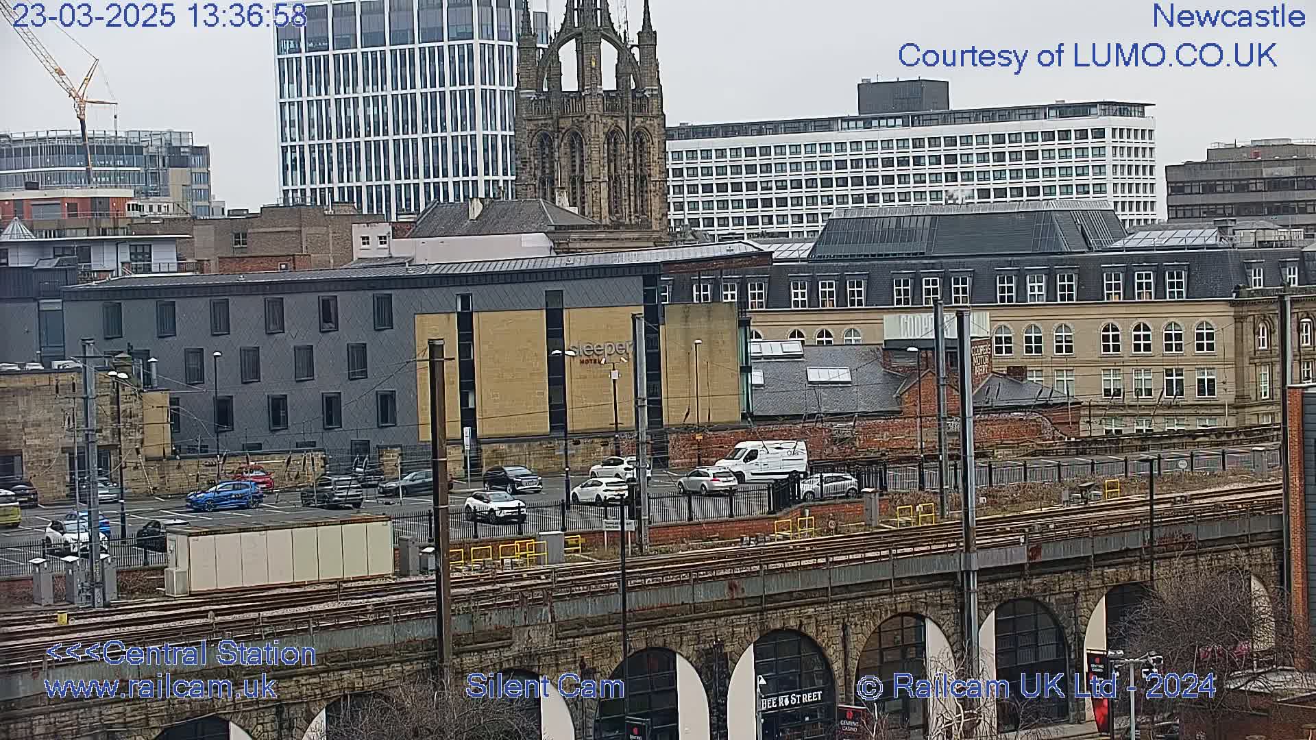 A city view showing a railway bridge over train tracks, a parking lot with cars, and various buildings including modern high rises and older stone structures, under an overcast sky.