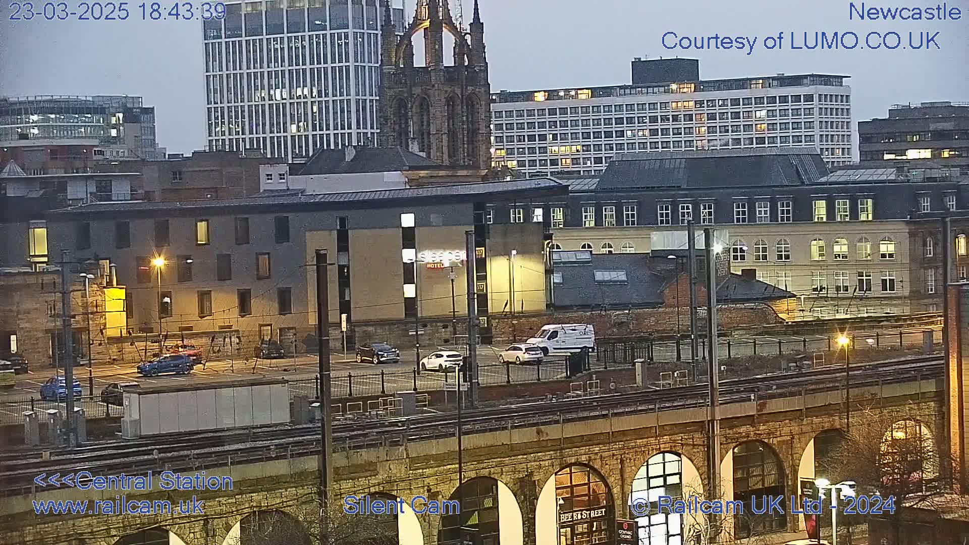 A nighttime view of a city featuring several buildings, a parking lot with cars, train tracks, and an arched stone structure, under a cloudy sky.