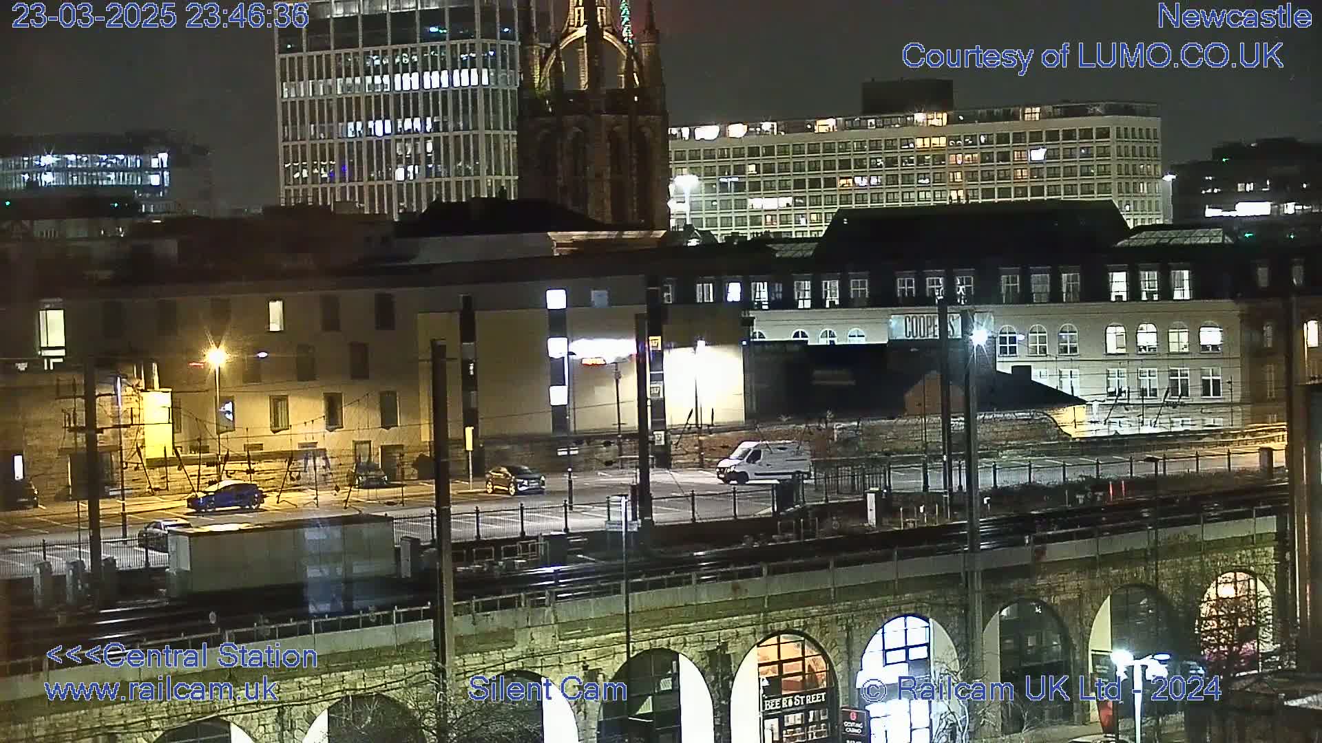 A nighttime view of a city with several buildings, a parking lot with vehicles, and a train bridge, under a dark sky.