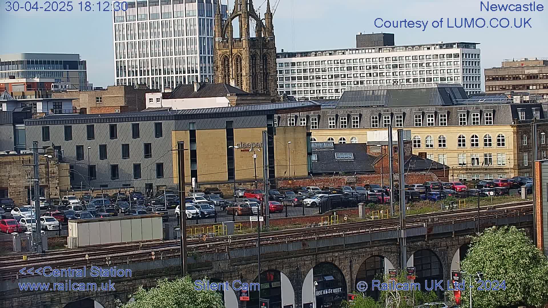 A city skyline view, under a clear sky, shows a large parking lot filled with cars near a train station and various buildings, including a church and a hotel.