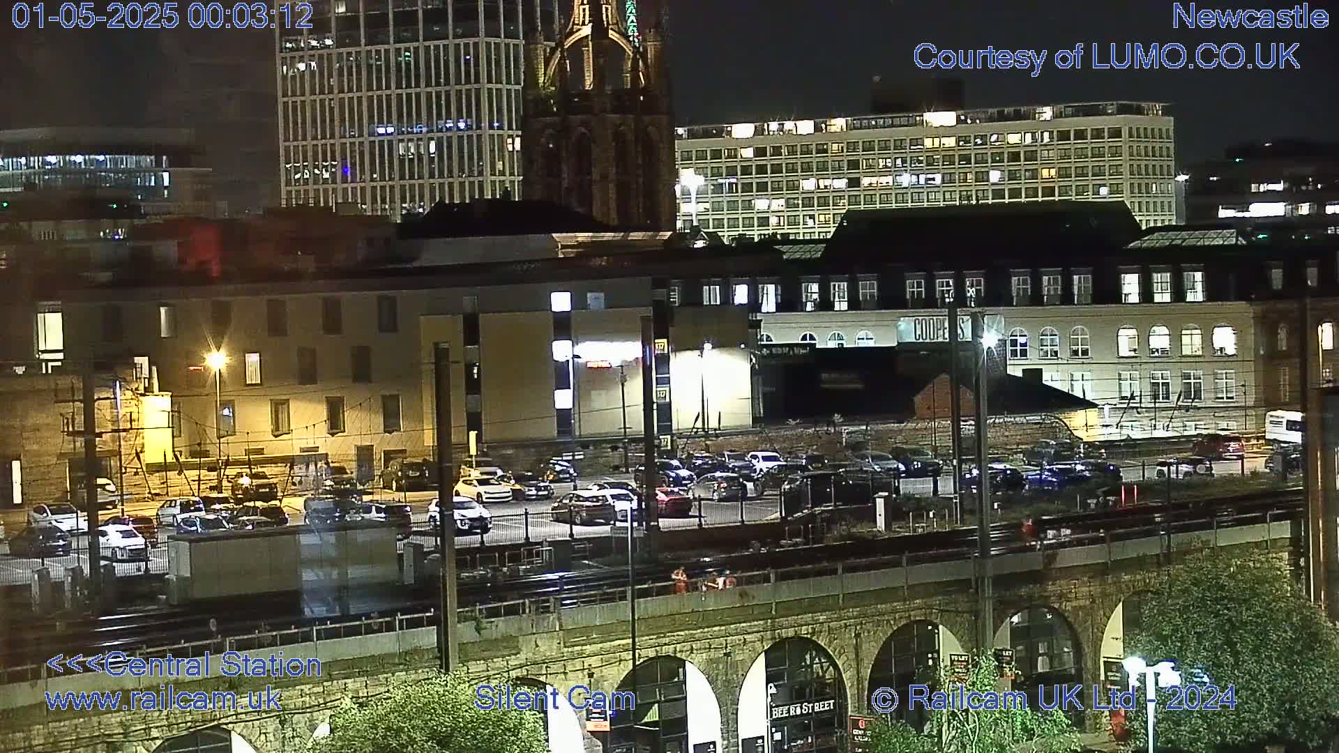 A nighttime view of a city featuring buildings, a parking lot full of cars, a train track, and a stone bridge.