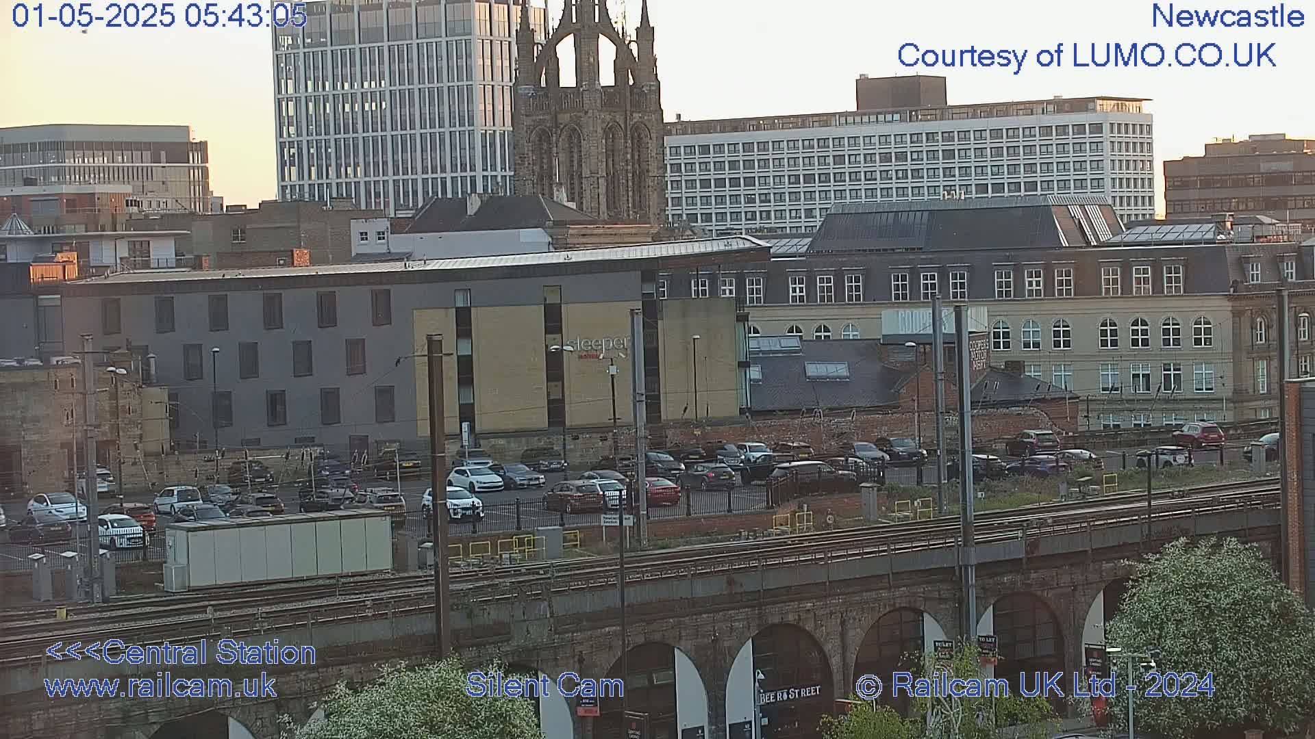 A city landscape at sunset, showing several buildings, a large parking lot filled with cars, and a train track below a stone archway.