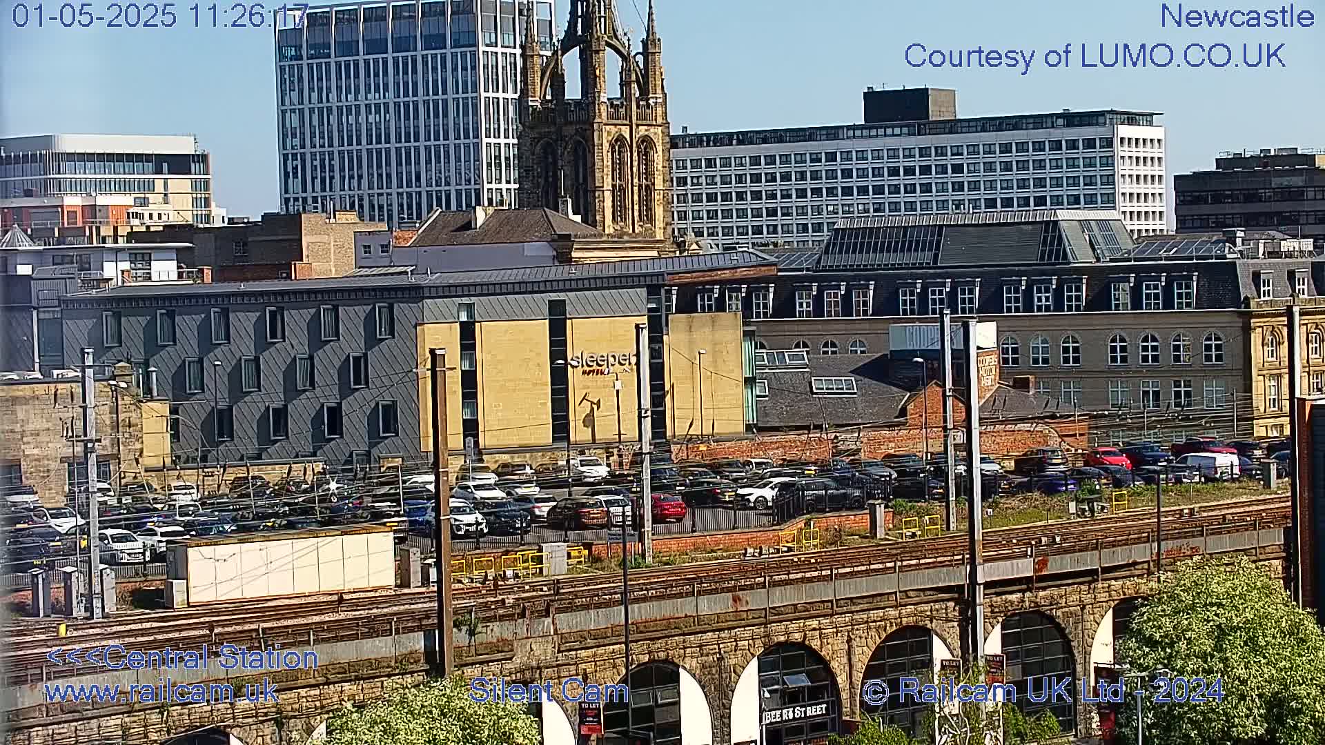 A sunny view of a city's skyline, featuring a large church, various buildings, a parking lot full of cars, and train tracks spanning a stone archway.