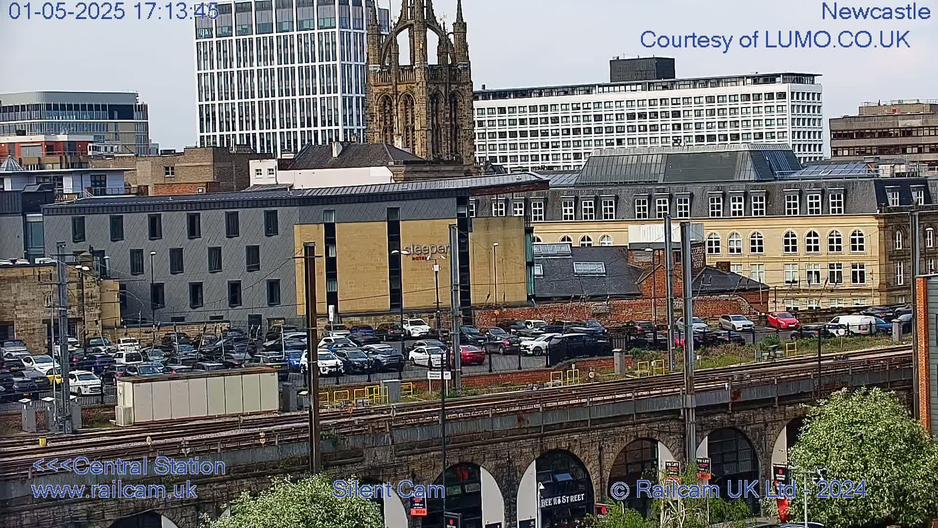 A city scene with a mix of modern and older buildings, a large parking lot, and train tracks viewed from an elevated position on a partly cloudy day.