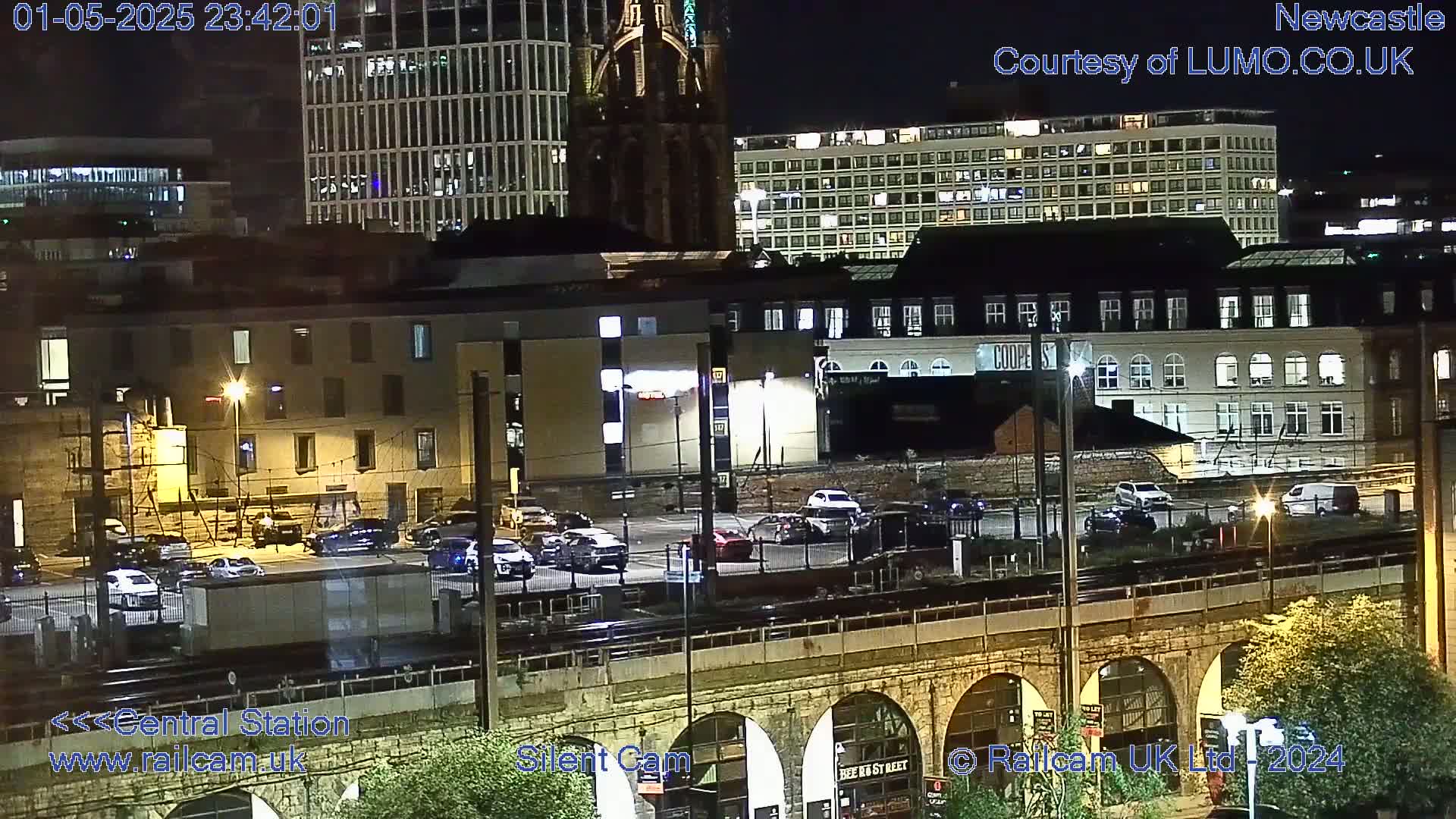 A nighttime view of a city's railway station area, with cars parked in lots and buildings illuminated.