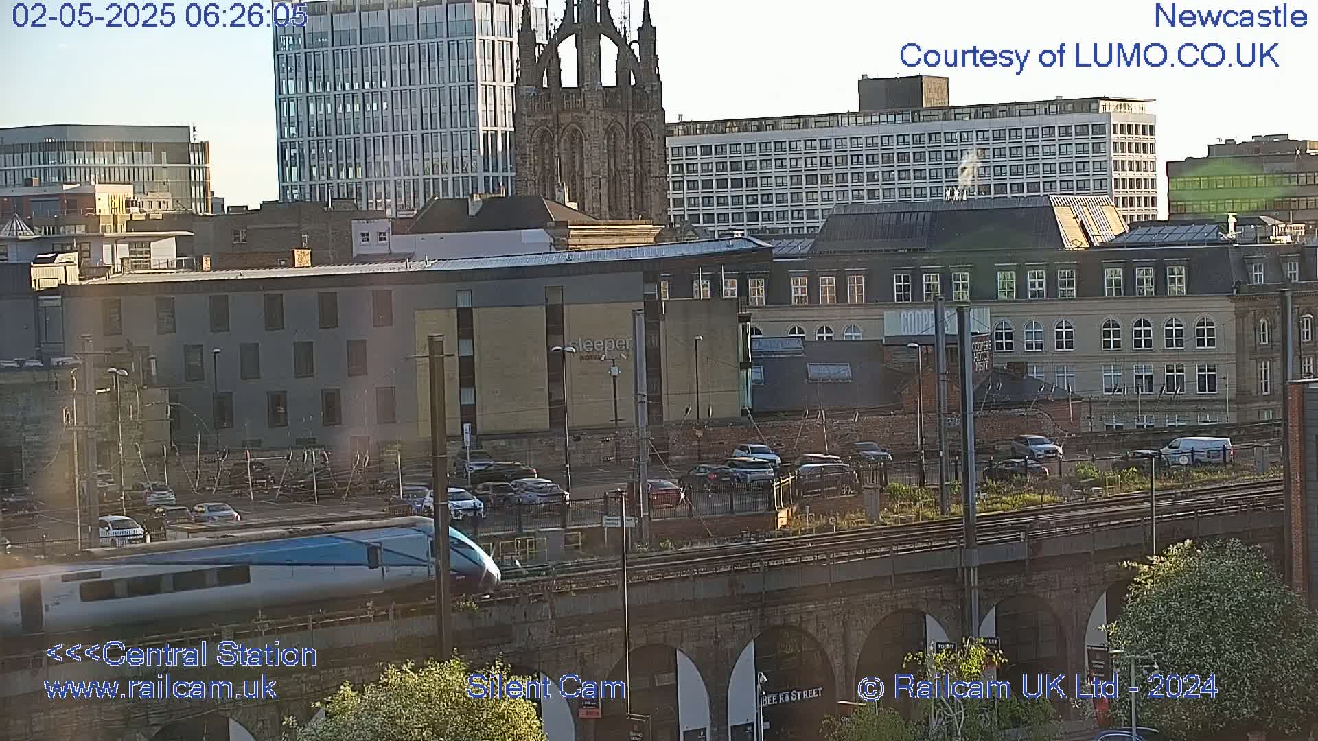 A high-speed train passes below a cityscape on a sunny day, with several buildings and parked cars visible in the background.