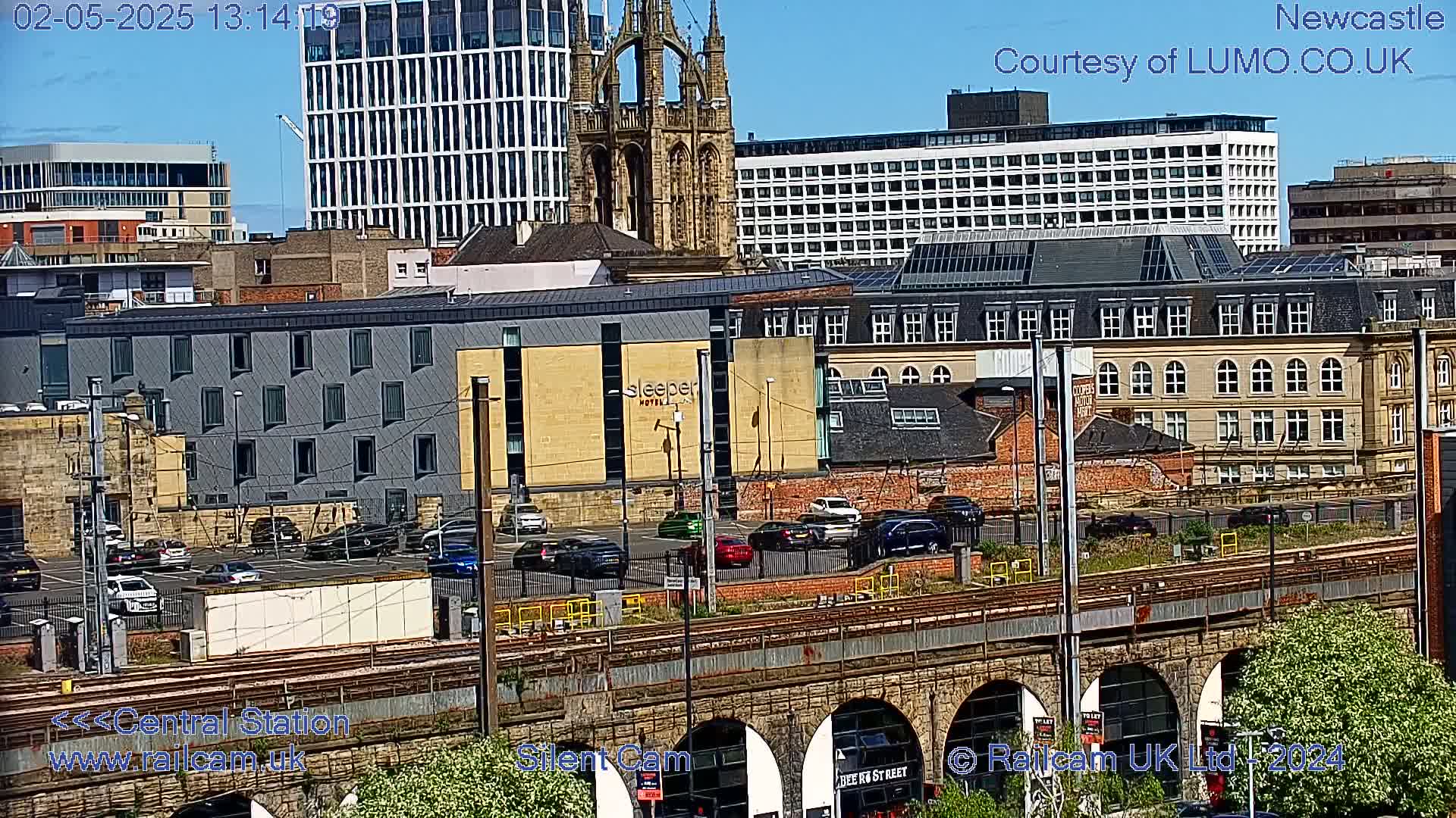 A sunny view of a city landscape shows various buildings, including a church, modern structures, and a hotel, with a railway and cars parked below, seen from an elevated vantage point.