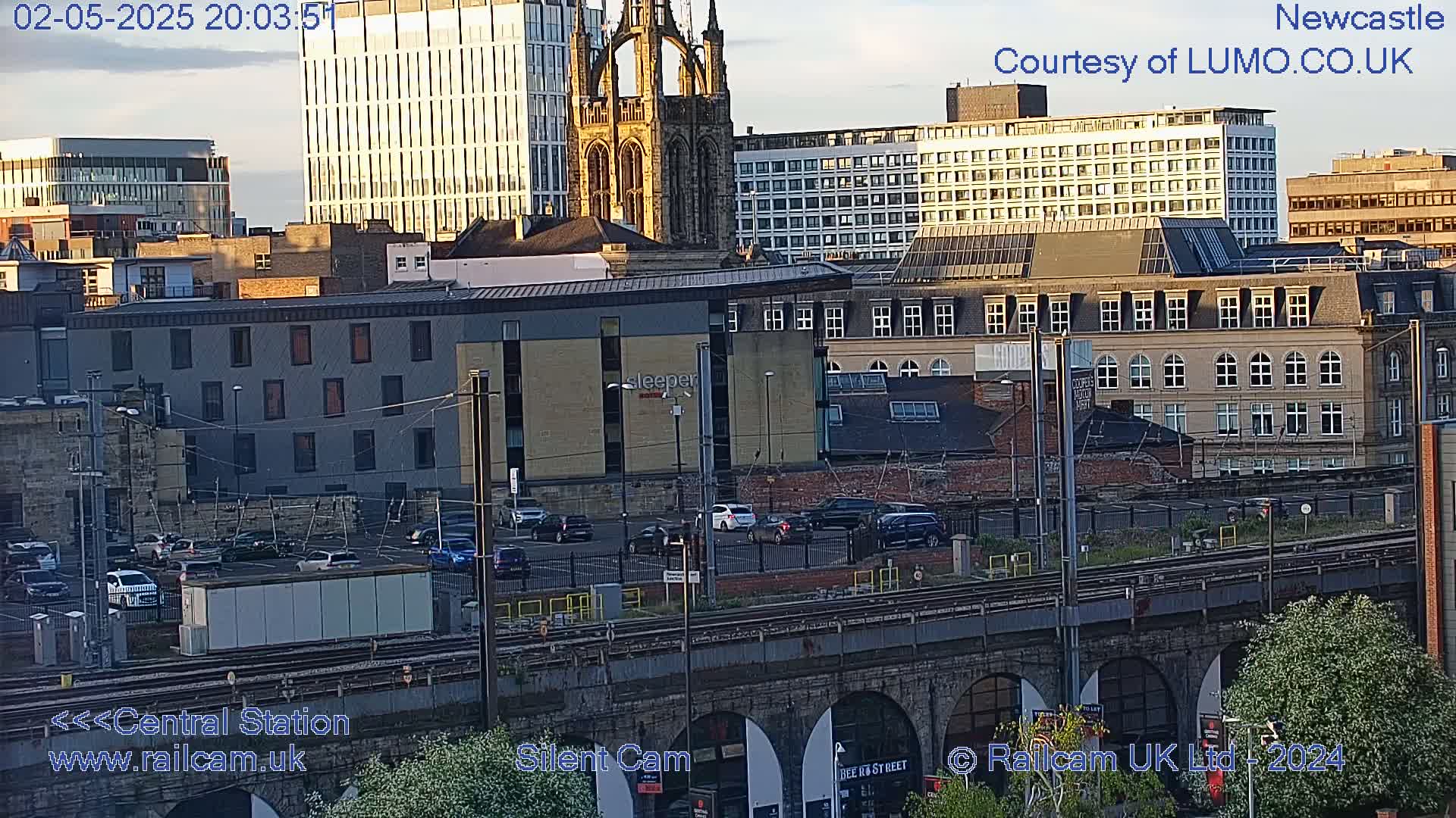 A city skyline at sunset, viewed from above a railway viaduct, showing various buildings including a tall church and modern high-rises.