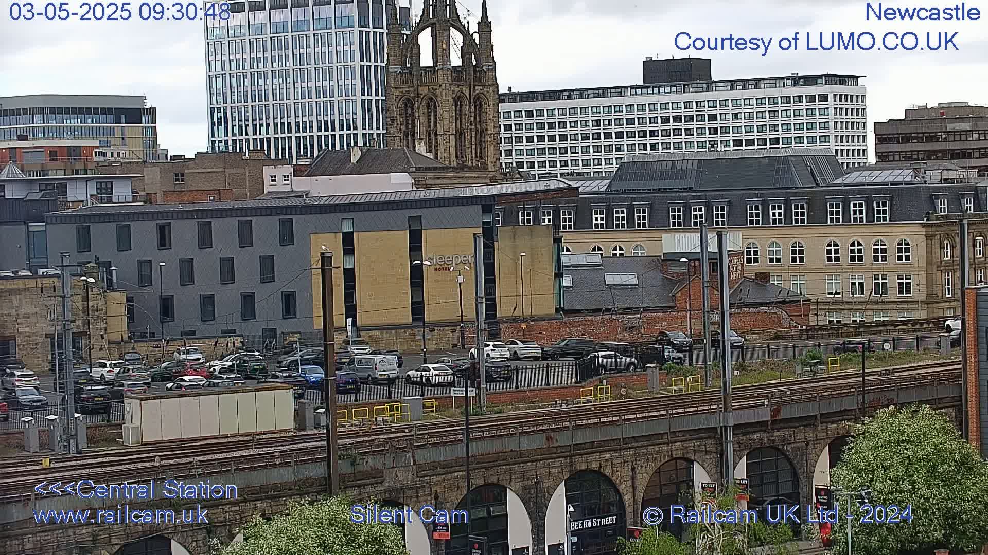A city skyline with numerous buildings, including a tall church and modern high-rises, is viewed from above a railway viaduct on an overcast day.