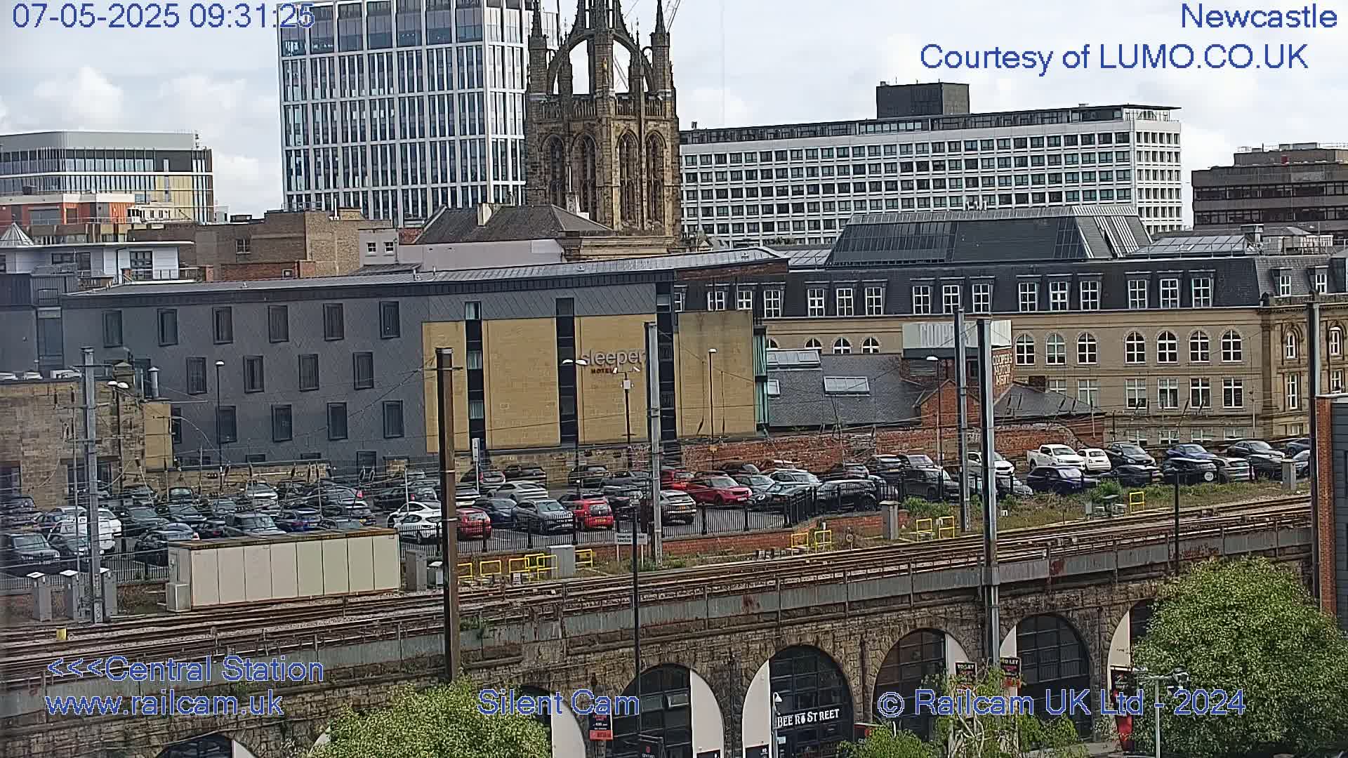 A city view under partly cloudy skies shows a large parking lot next to train tracks, with various buildings including a tall modern building, several multi-story buildings, and a church-like structure in the background.