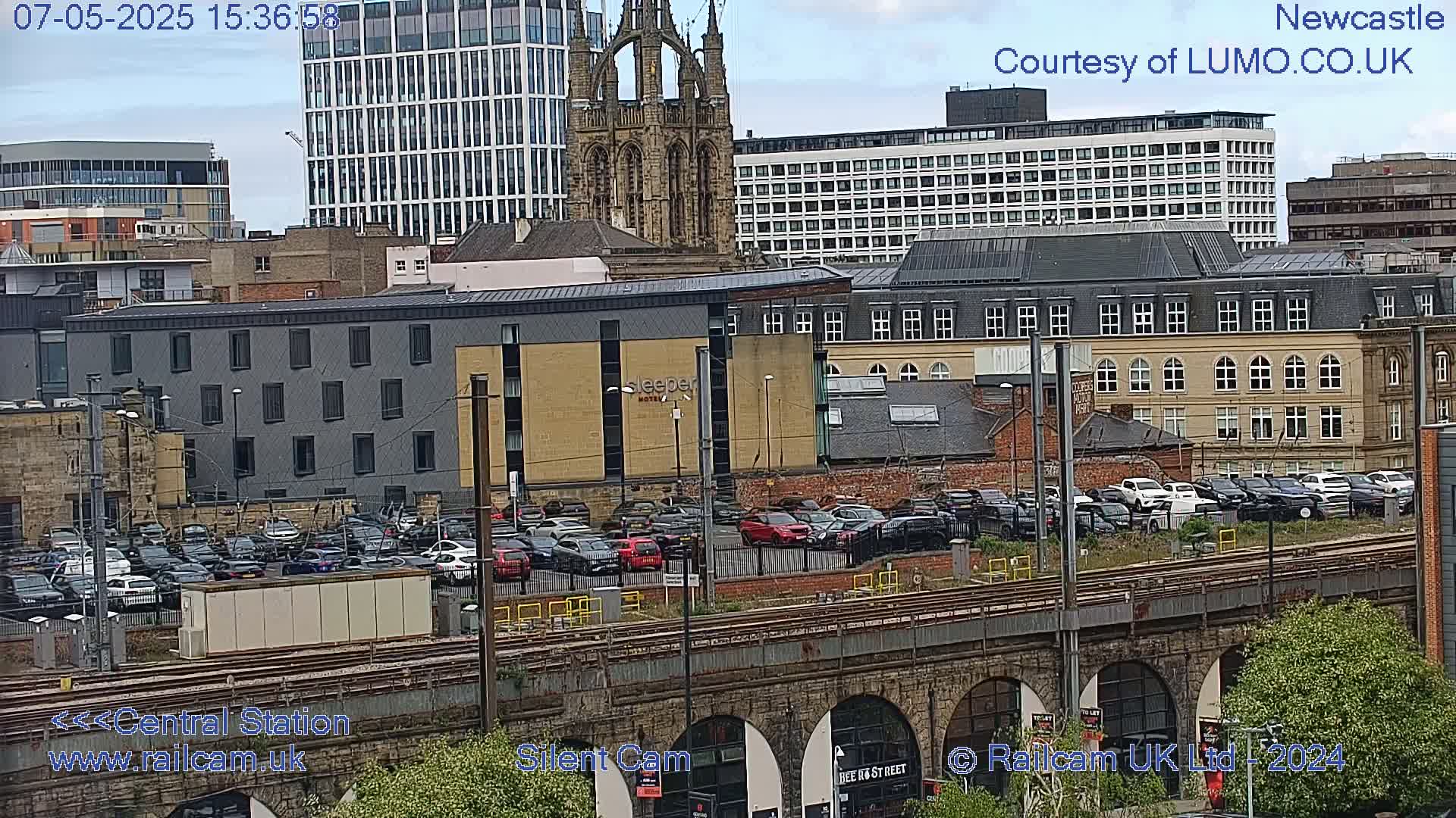 A city scene under an overcast sky shows a large parking lot full of cars next to train tracks and a stone bridge, with various buildings, including a church and modern high-rises, in the background.
