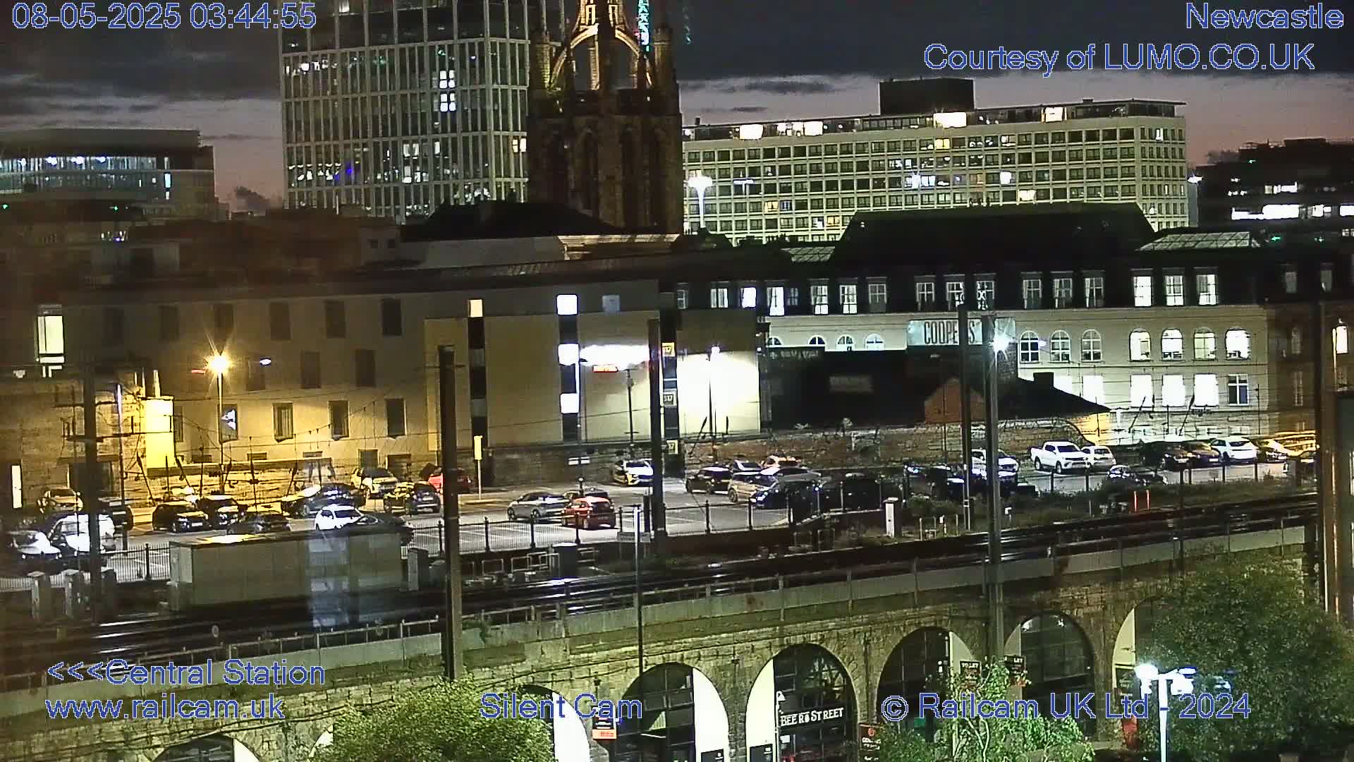 A nighttime view of a city's central station area, with a parking lot, train tracks, and a stone arch bridge, under a partly cloudy sky.