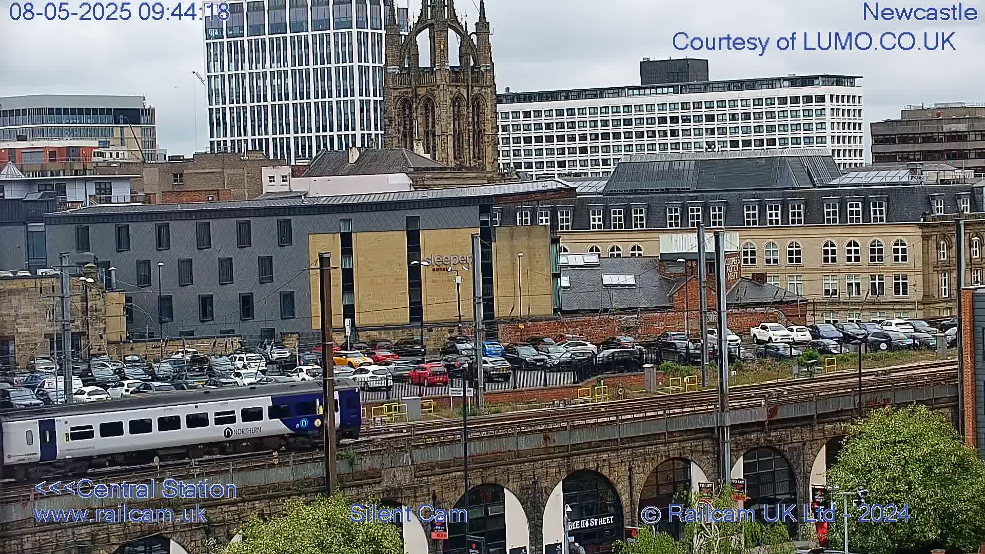 A train passes by a city's railway tracks, under an overcast sky, with various buildings and parked cars visible in the background.