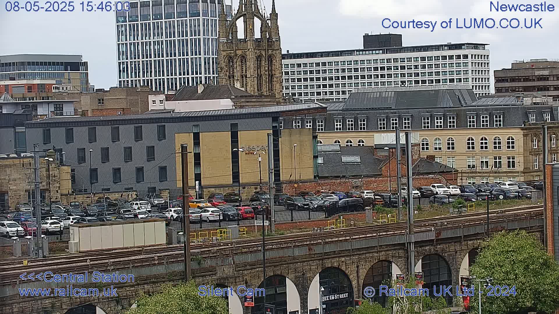 A city landscape with overcast skies shows a parking lot filled with cars next to a train track and several buildings, including a tall modern building and a church.