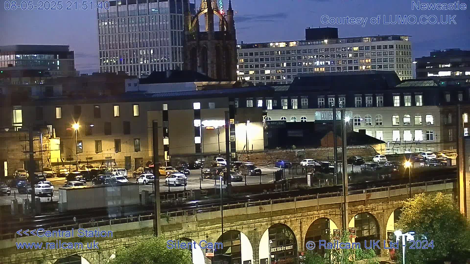 A nighttime view of a city's central station area, showing several buildings, a parking lot with cars, and a stone arch bridge, under a twilight sky.