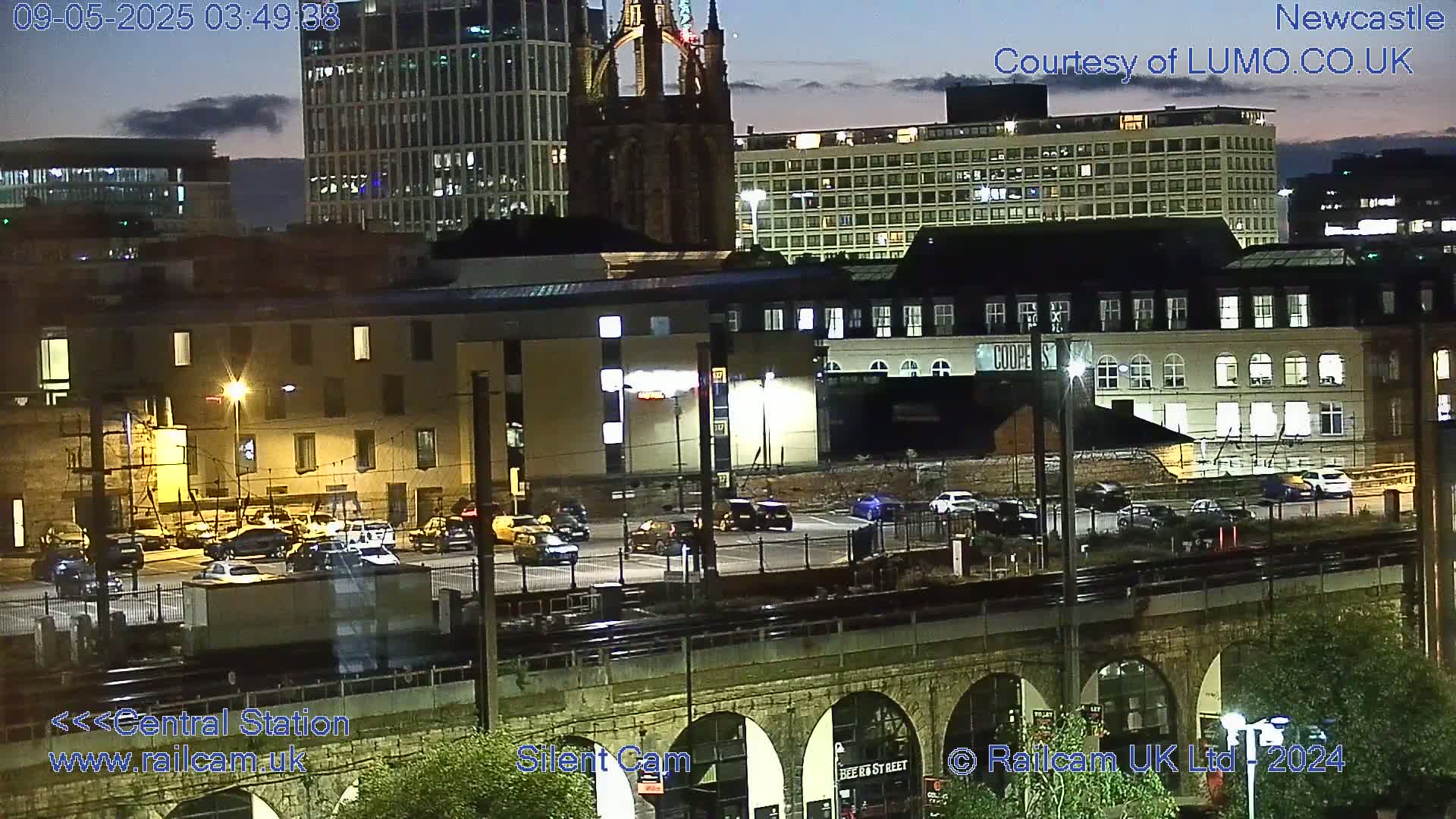 A nighttime view of a city featuring several buildings, a parking lot with cars, and a train track passing under an arched stone bridge.