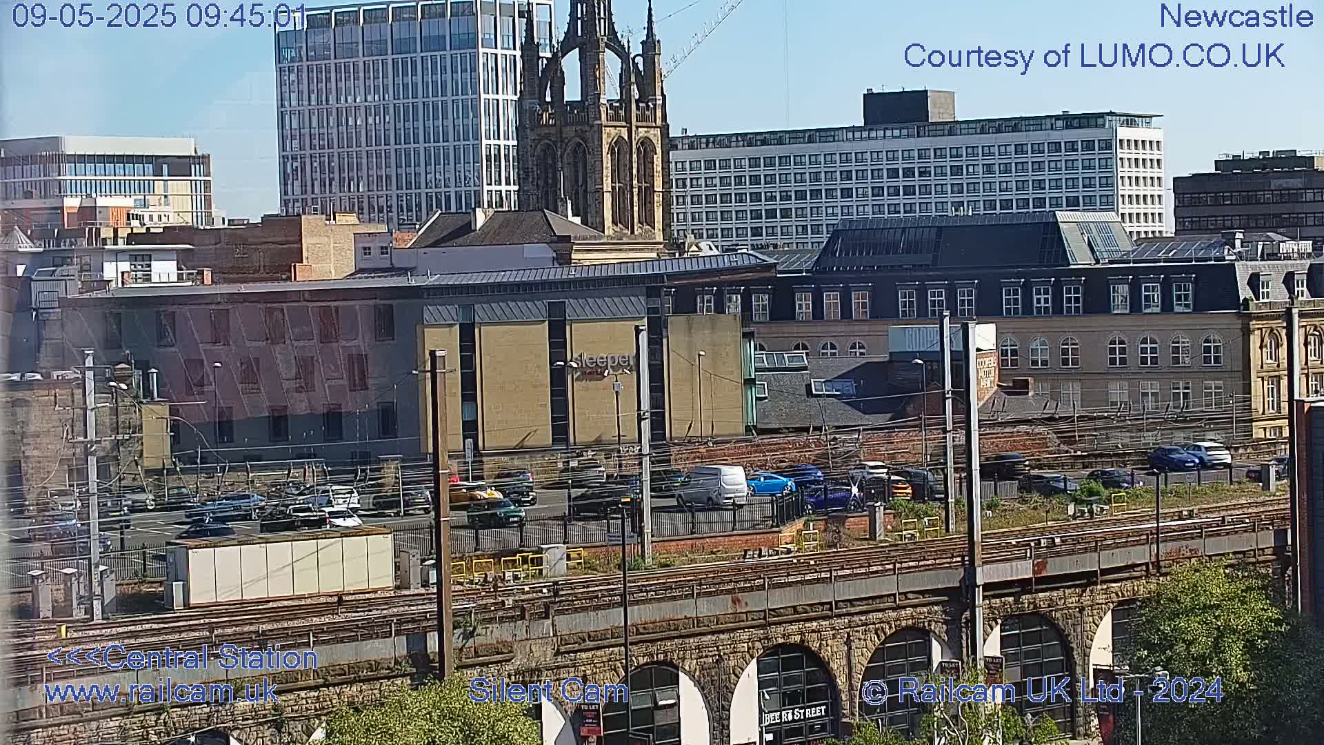 A city skyline with a church steeple, modern and older buildings, a train station, and cars, under a sunny sky.