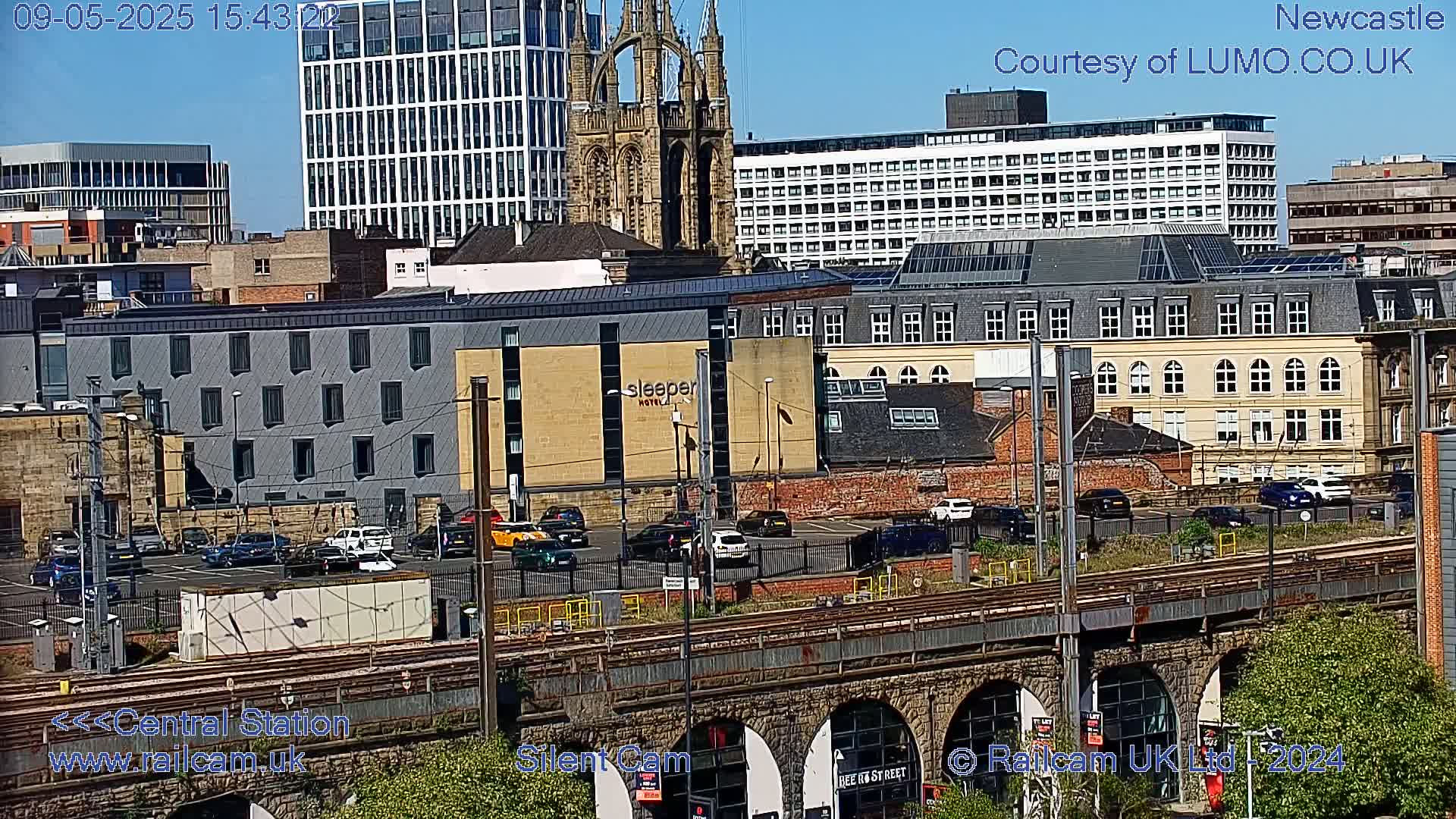 A sunny city view shows a railway viaduct above a parking lot and several buildings, including a tall modern building and a gothic-style church, with a hotel prominently displayed.