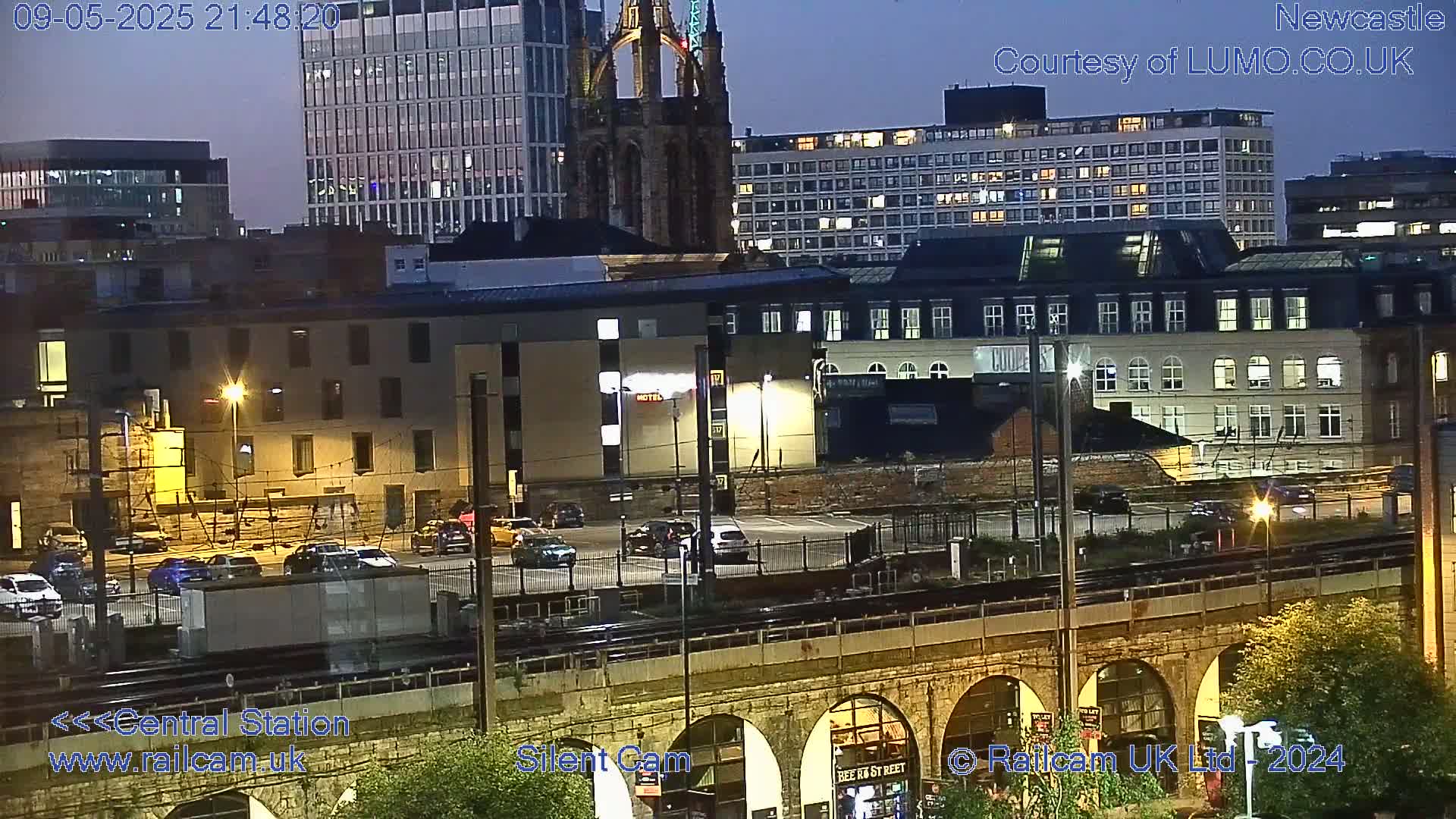 A nighttime view of a city's cityscape, featuring buildings, a parking lot with cars, and a railway bridge under a twilight sky.