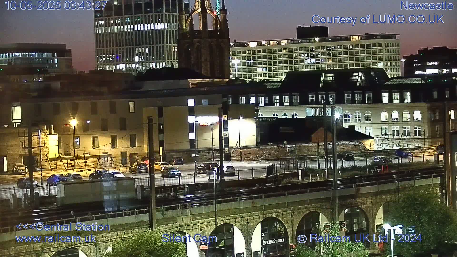 A nighttime view of a city’s illuminated buildings and a railway station, seen from above a stone arch bridge.