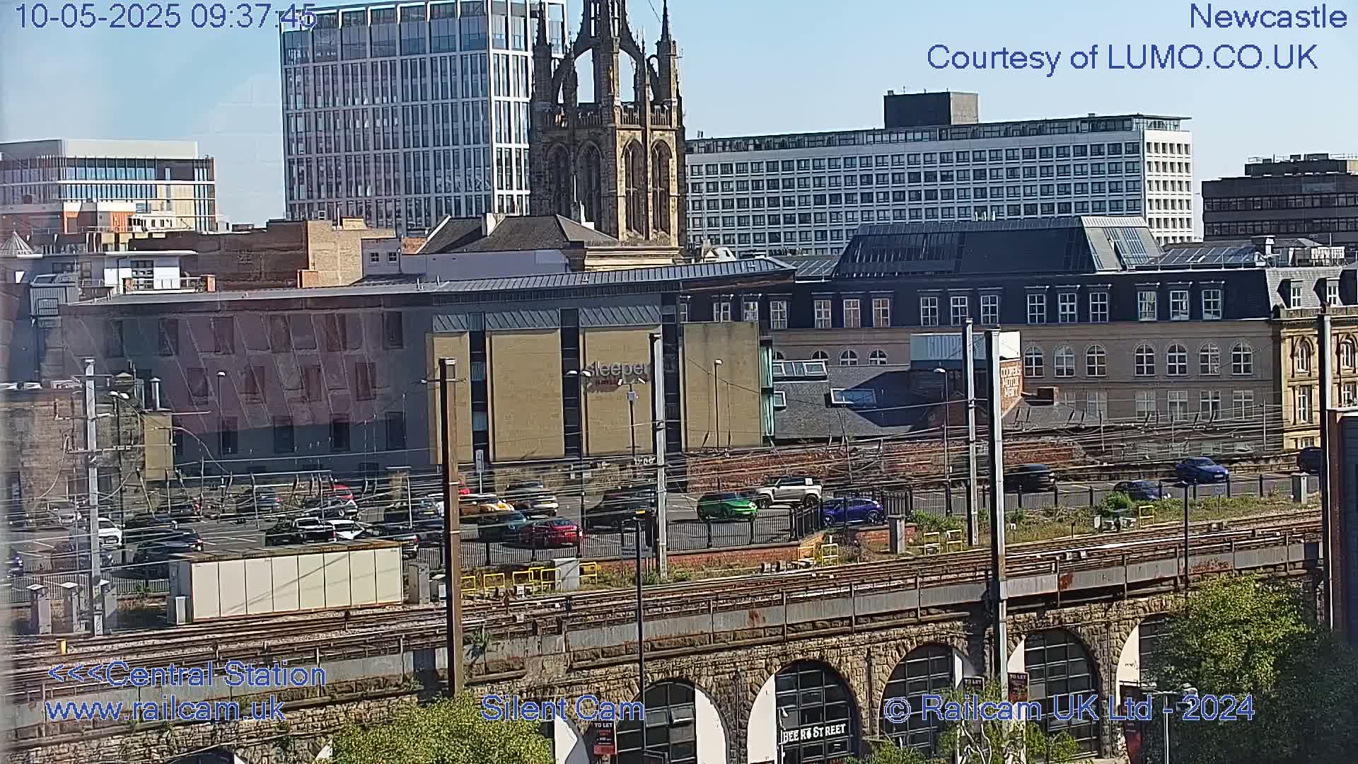 A sunny cityscape view shows numerous buildings, a railway, and parked cars, with a tall church spire visible in the mid-ground.