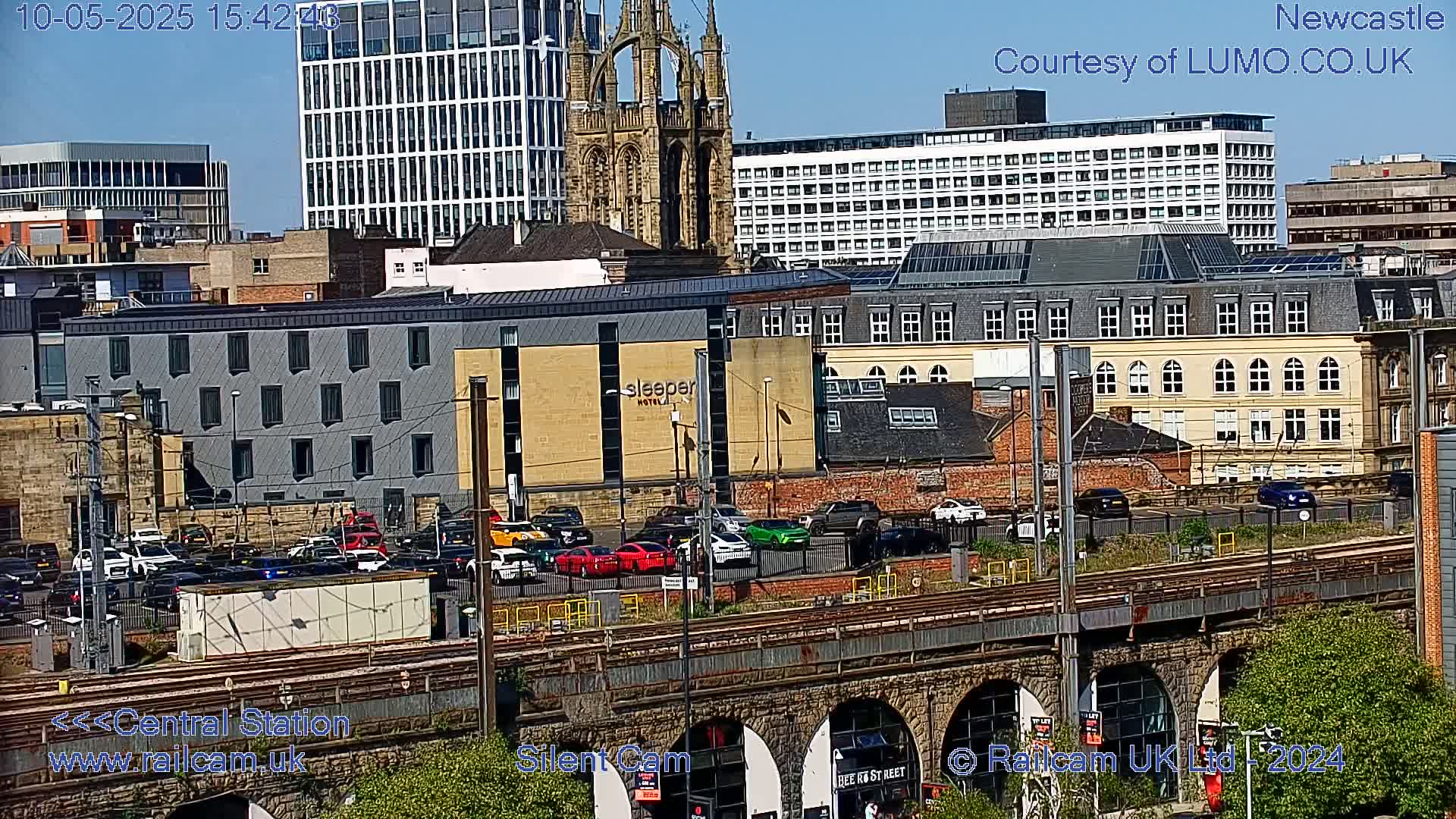 A sunny view of a city cityscape with several buildings, a railway line, and a significant amount of traffic, including cars parked and driving.
