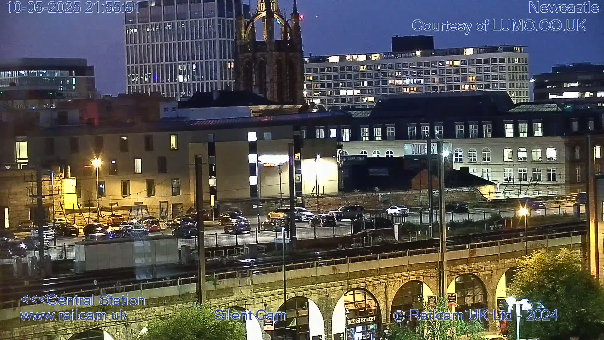 A nighttime city scene viewed from above shows a railway viaduct, parked cars in a lot, and illuminated buildings including a church and high-rises.