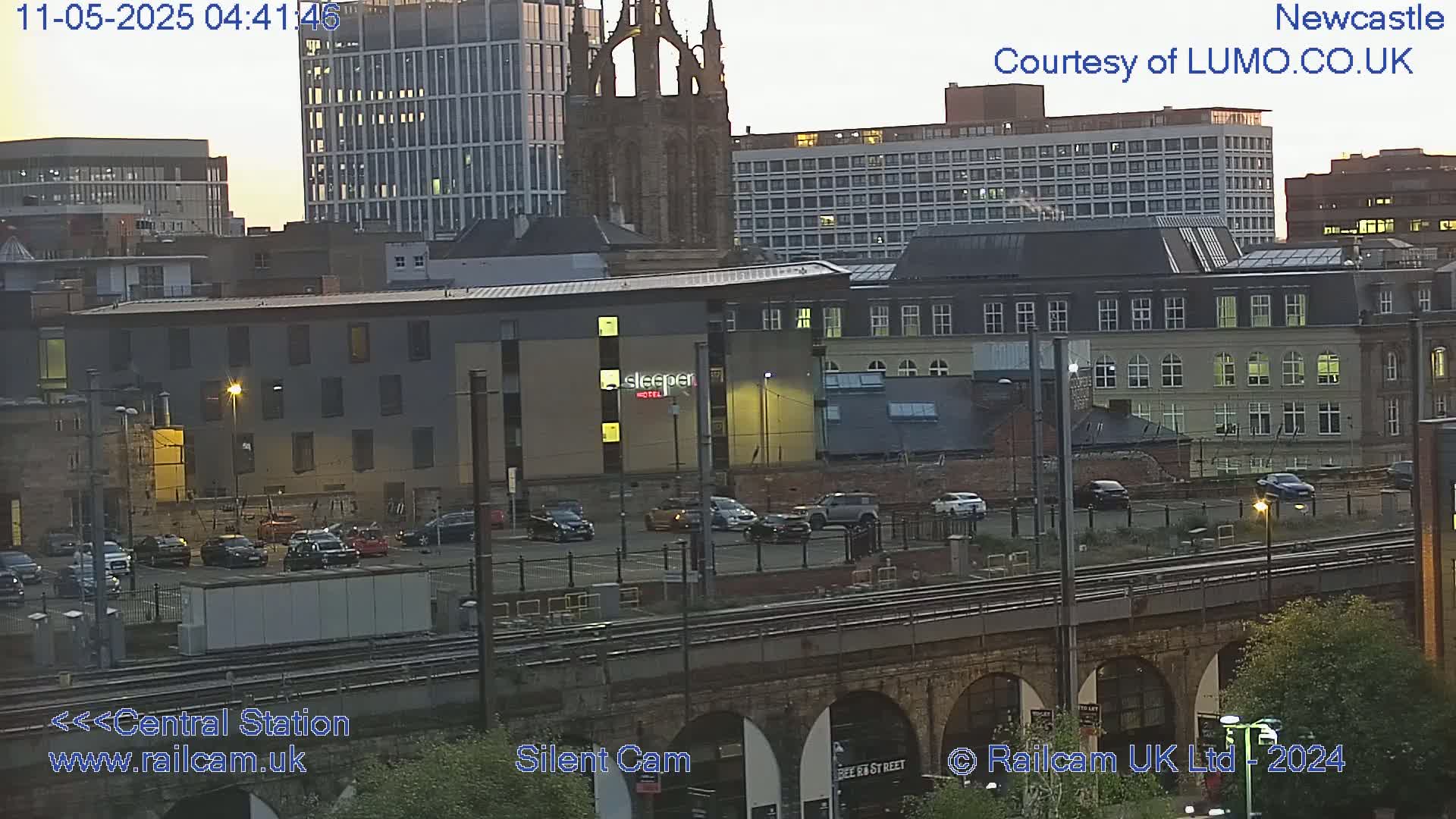 A city scene at twilight shows a parking lot filled with cars in front of several buildings, including a hotel, next to a railway viaduct.