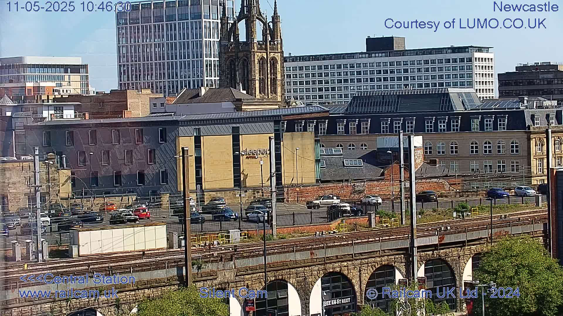 A sunny view of a city with various buildings, including a tall church, a hotel, and other structures, seen from above a train track and stone arches.