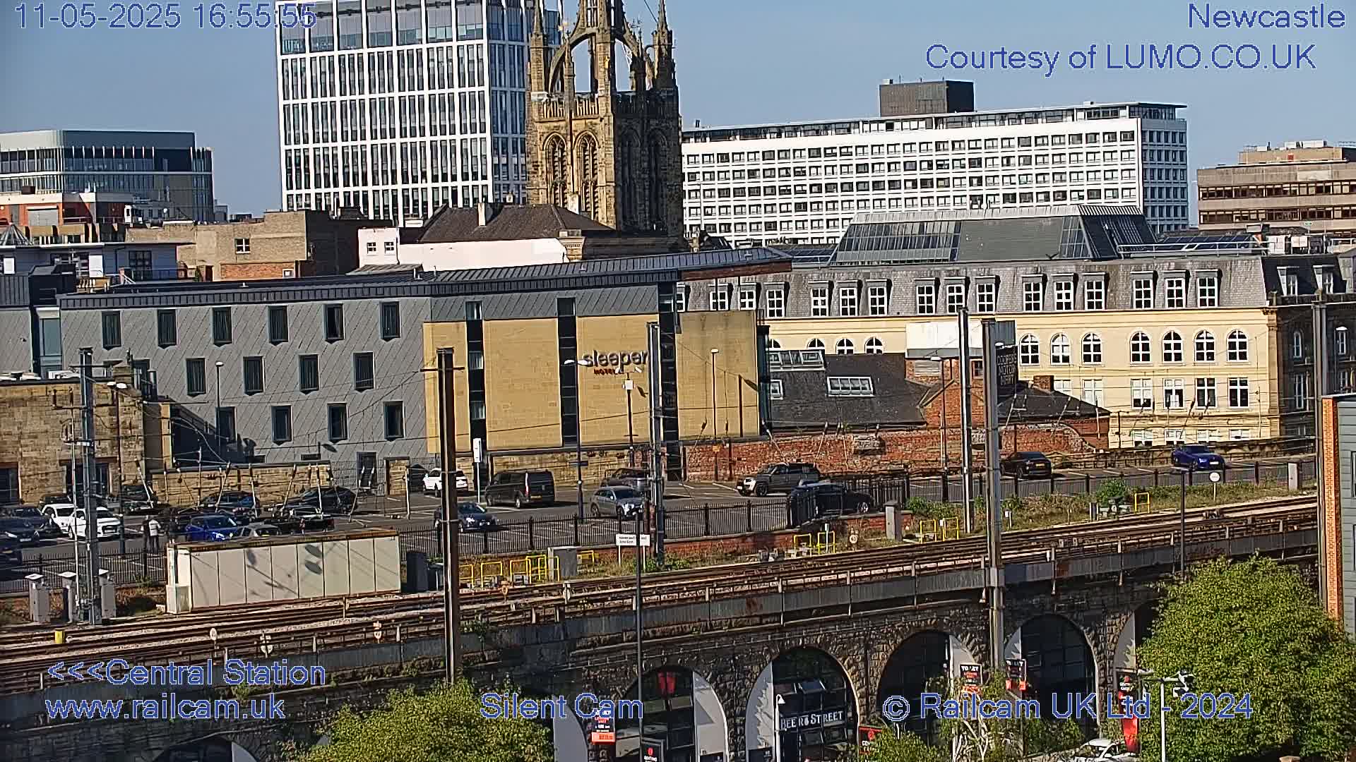 A city view on a sunny day shows several buildings, including a church, a parking lot with cars, train tracks, and a stone archway.