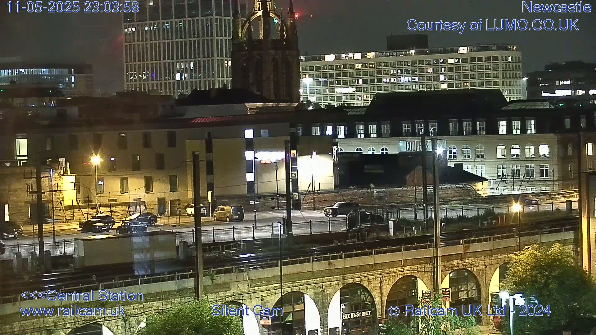 A nighttime view of a city with several buildings, vehicles in a parking lot, and a train track below an arched bridge, under a clear sky.
