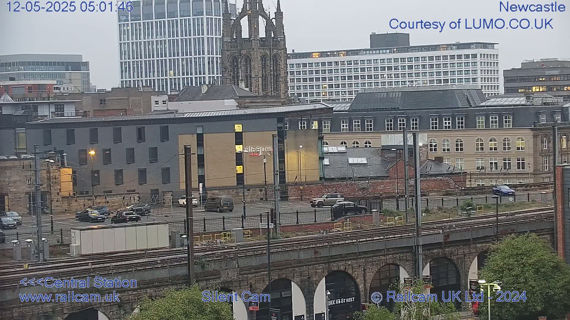 A view of a city including a parking lot, railway tracks, several buildings, and a church spire under an overcast sky.