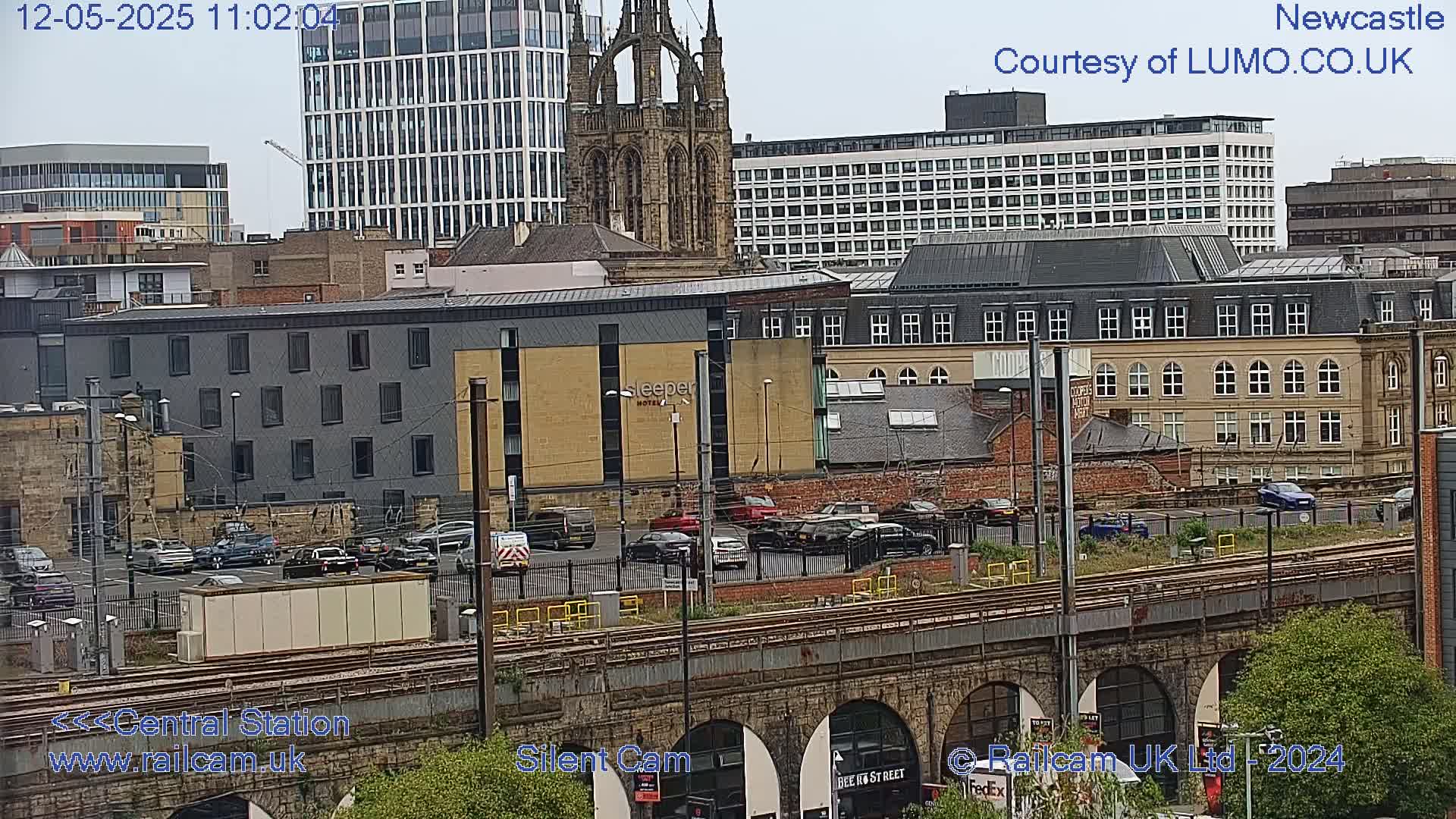 A city view from a bridge overlooking a train track and parking lot, with several buildings including a tall modern building and a Gothic-style church, on a partly cloudy day.