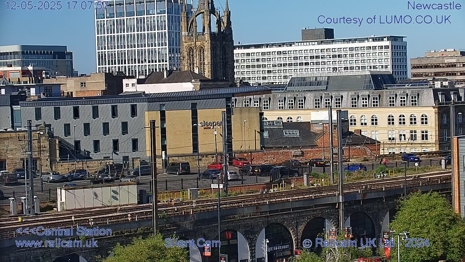 A city scene on a sunny day, featuring a variety of buildings including a tall church, modern high-rises, and smaller structures, with railway tracks and a bridge in the foreground, and cars parked in a lot.