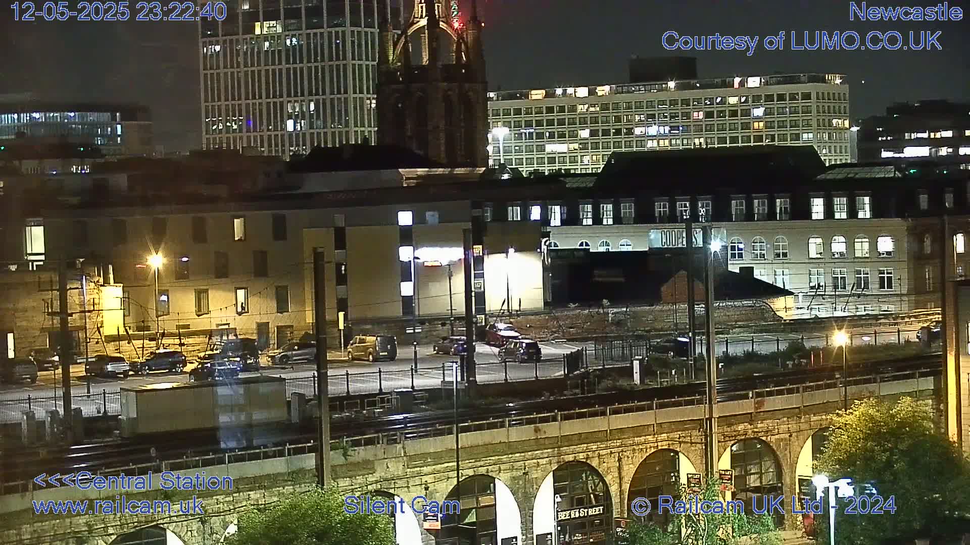 A nighttime city scene shows several buildings, a parking lot with cars, train tracks, and a stone archway bridge, illuminated by streetlights.