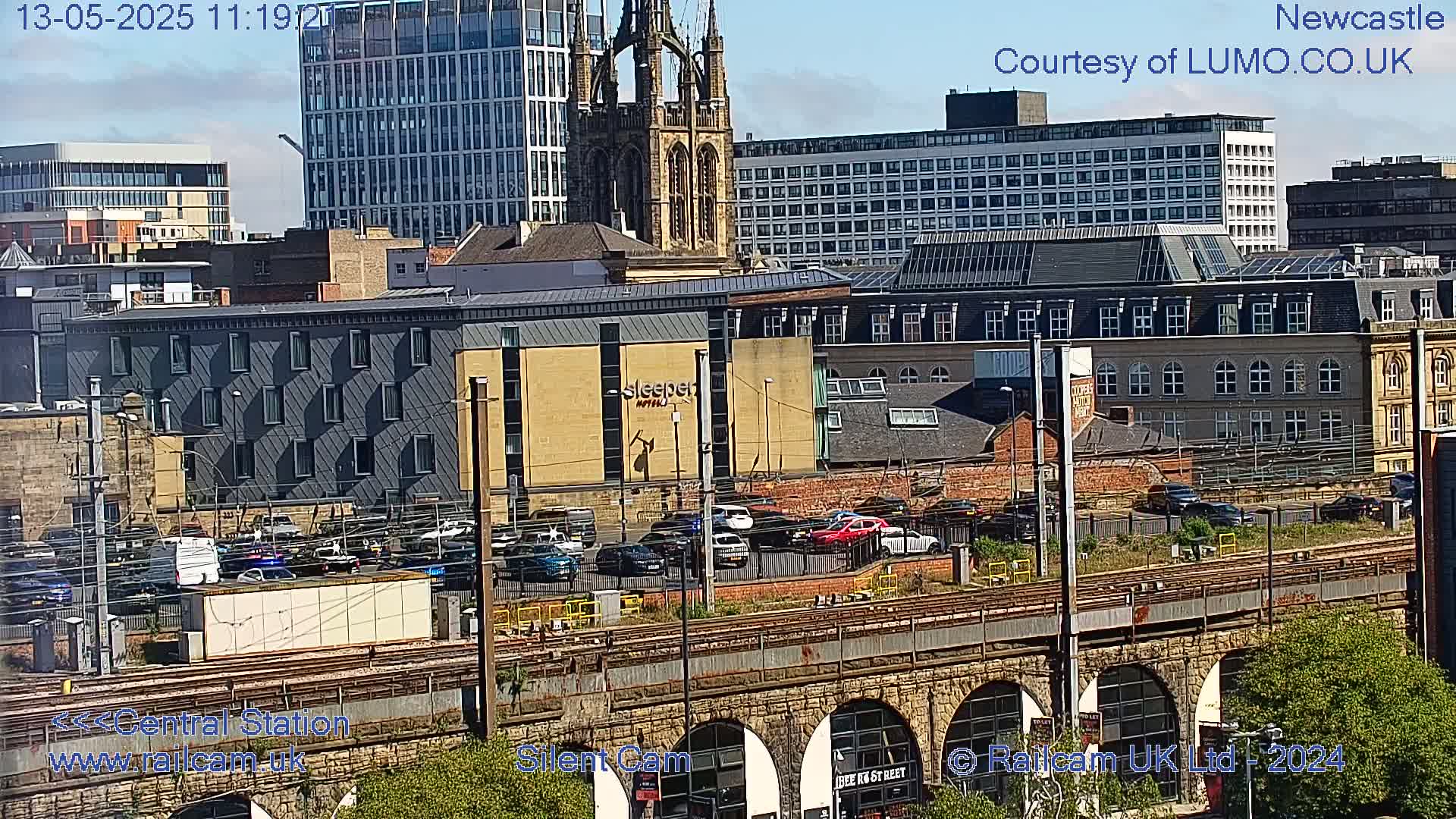 A sunny city scene viewed from above shows a railway viaduct, a parking lot with several cars, and a variety of buildings including a tall church and a modern hotel.