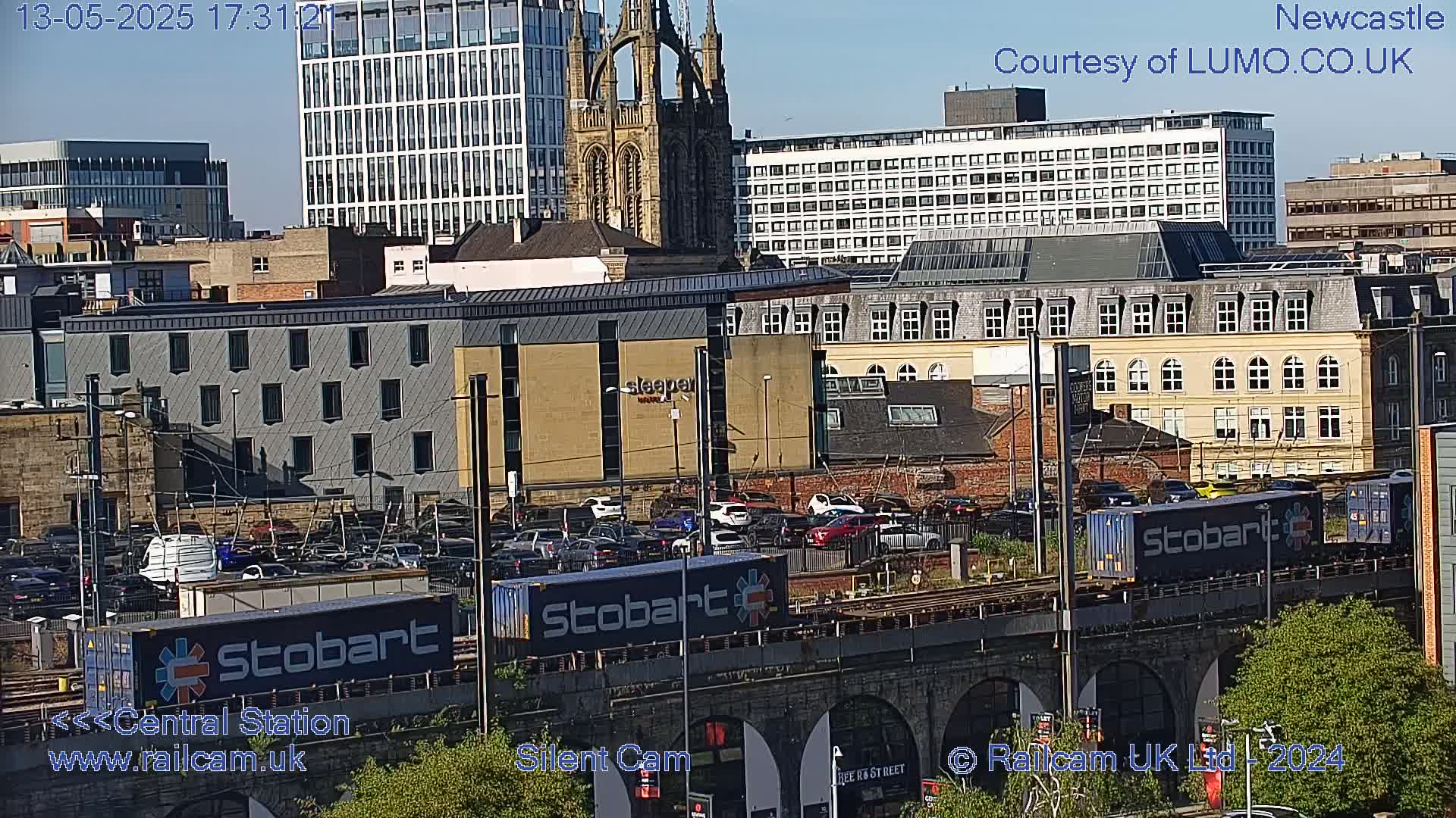 A sunny cityscape view shows a train carrying several Stobart containers passing over a stone bridge, with various buildings and a church in the background.