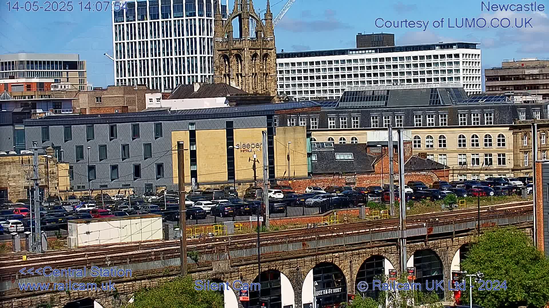 A city scene on a sunny day shows a large parking lot full of cars next to a train track that runs under a stone arch bridge, with various buildings, including a hotel and a church, in the background.