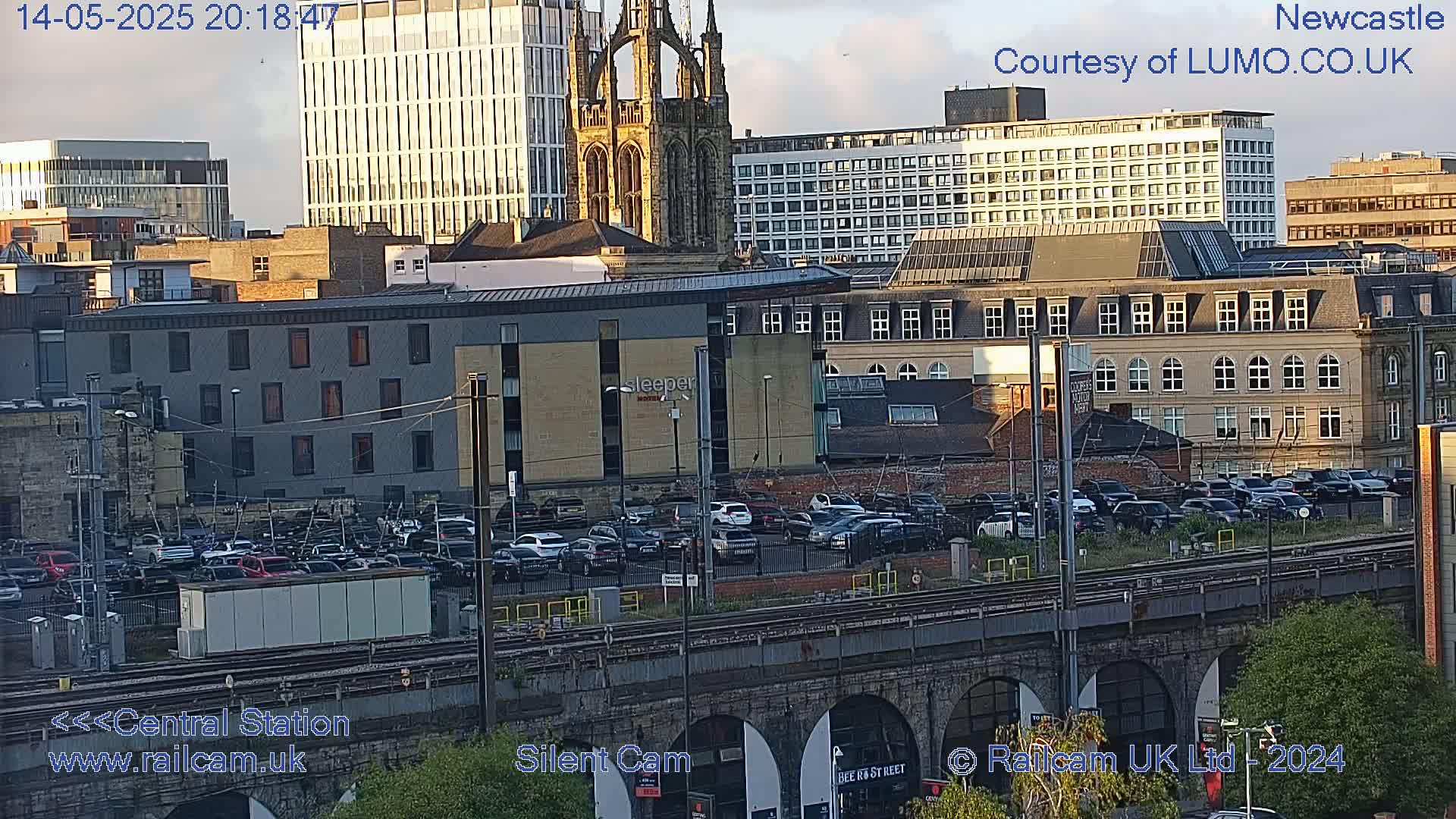 A city skyline at sunset, showing a mix of modern and older buildings, with a large parking lot and railway tracks in the foreground.
