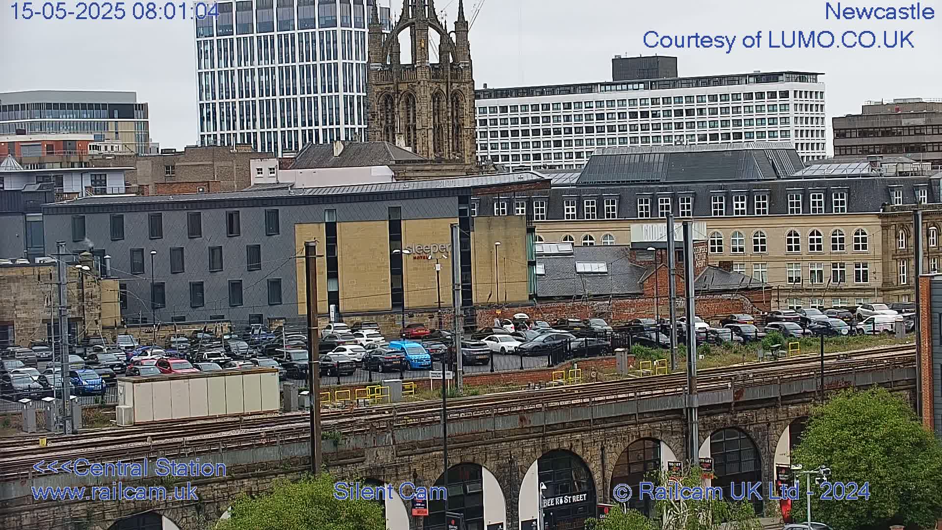 A city view on an overcast day shows a large parking lot filled with cars next to a train track and bridge, with various buildings and a church spire in the background.