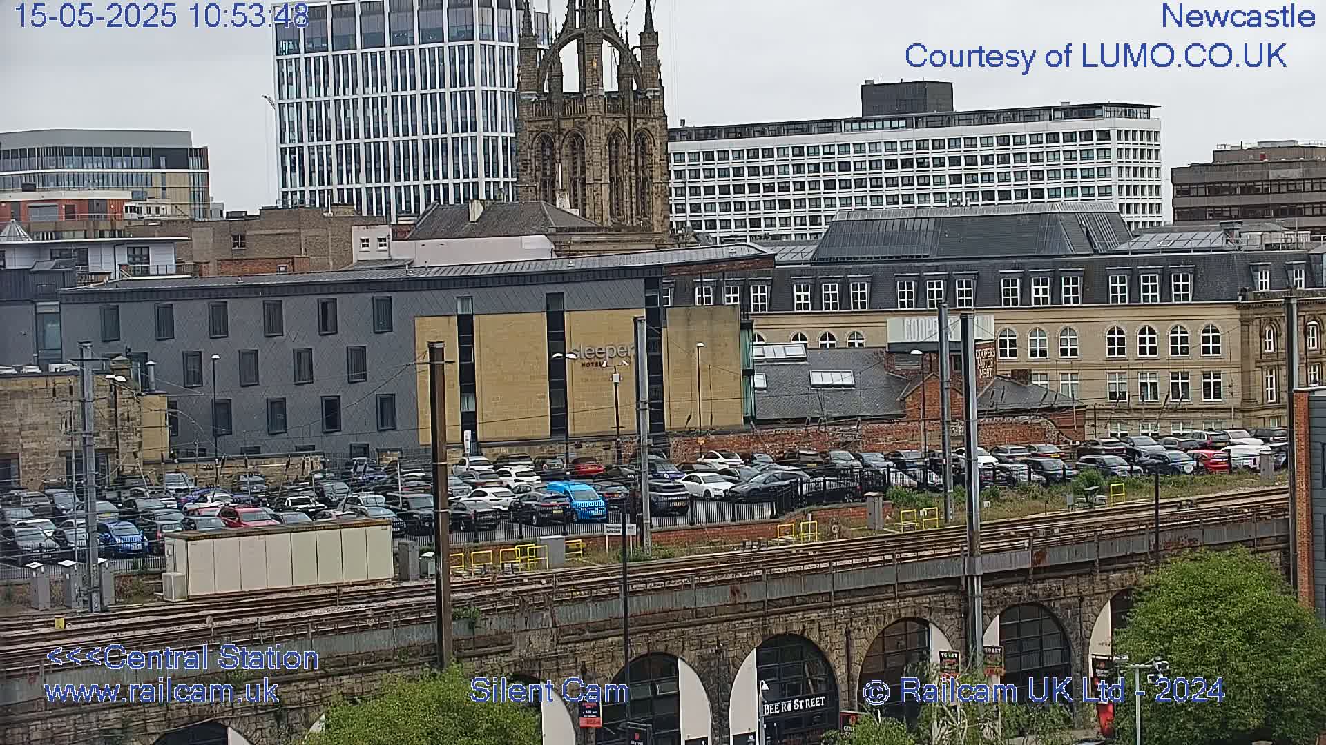 A large parking lot filled with cars sits beside a railway, with various buildings, including a tall modern structure and a church, in the background under an overcast sky.