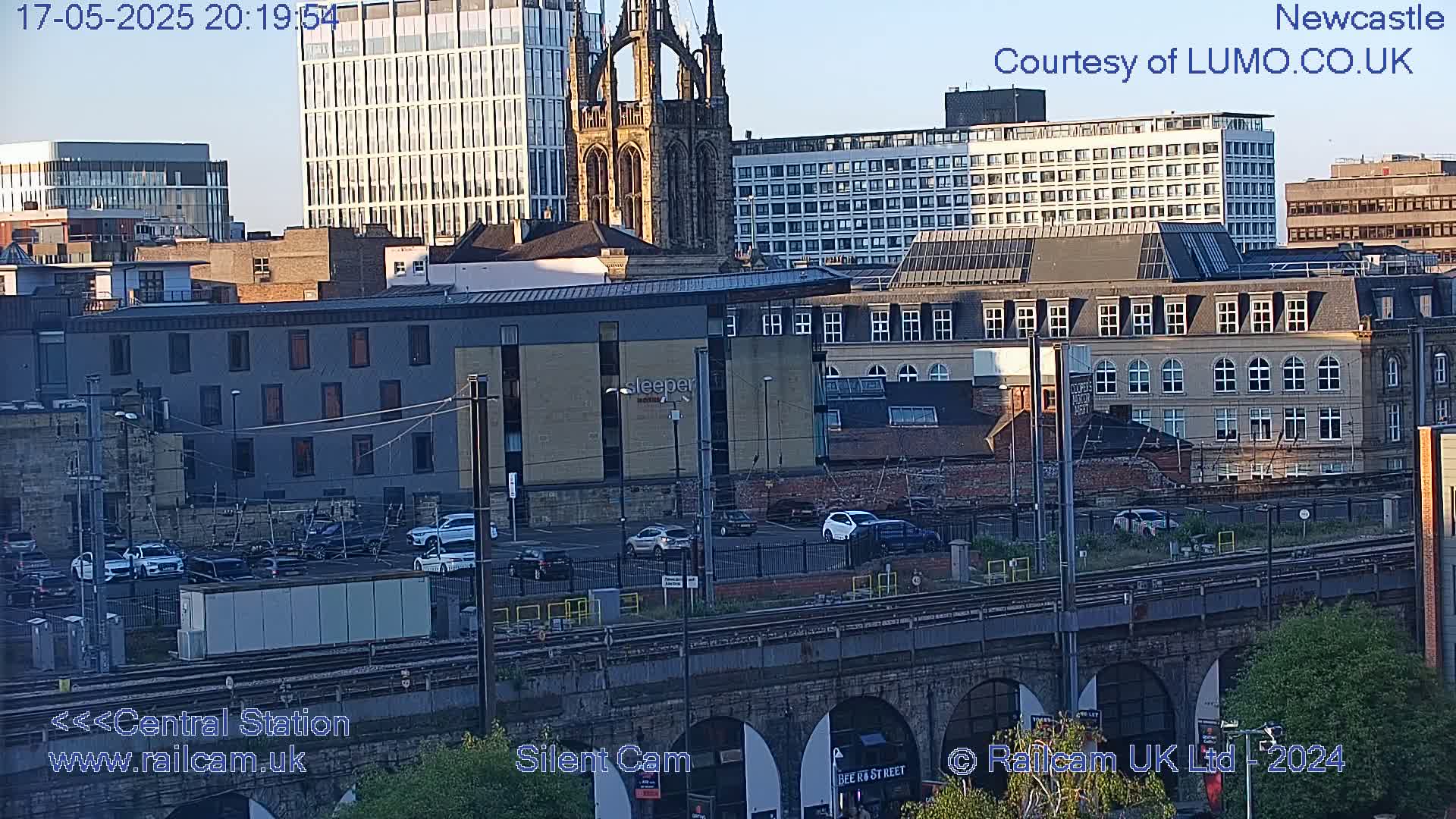 A view of a city skyline at dusk, featuring several buildings, a railway, and cars parked in a lot.
