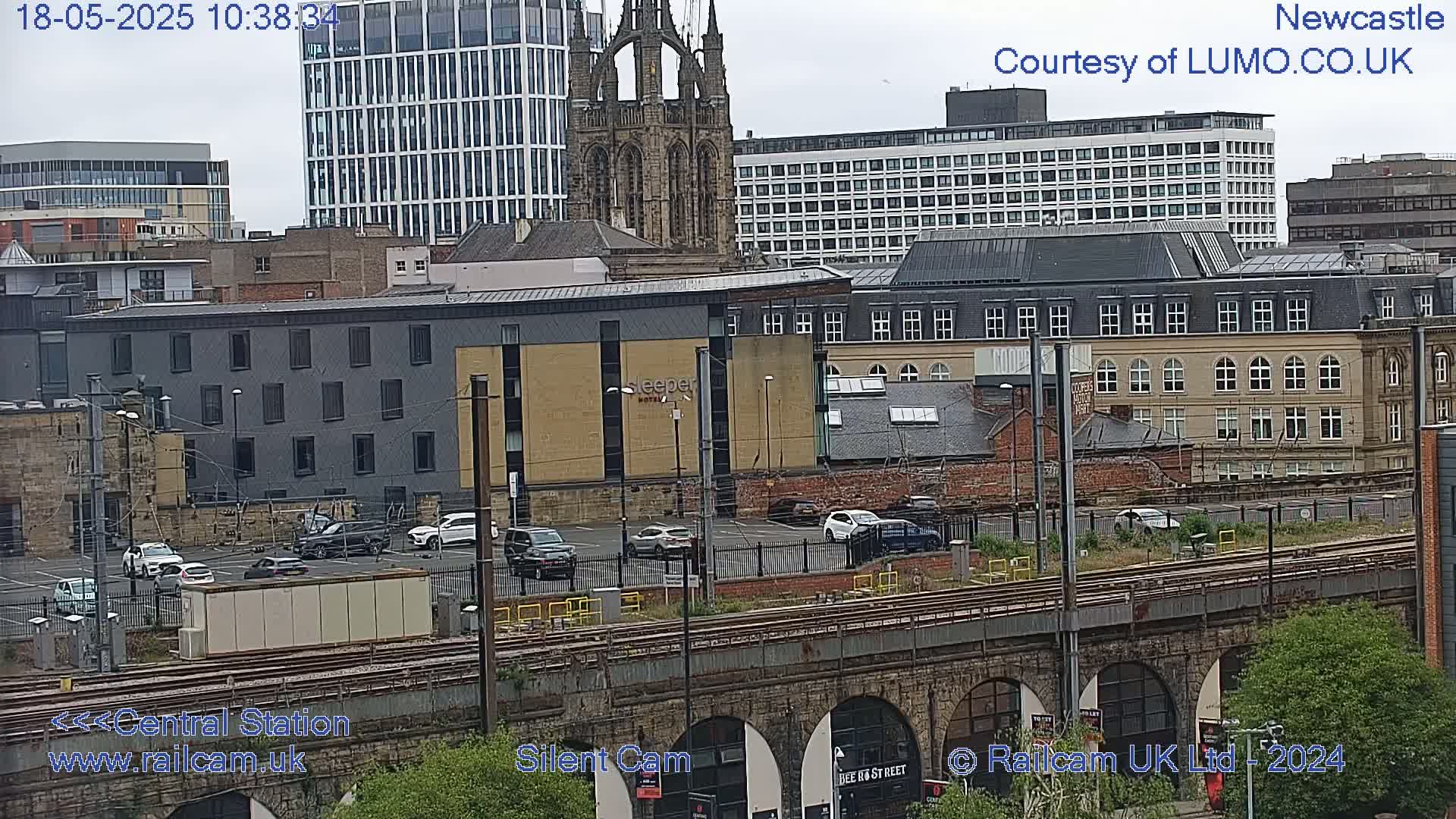 A city scene viewed from above on an overcast day, showing a parking lot, railway tracks, and various buildings including a tall modern structure and a Gothic-style church.